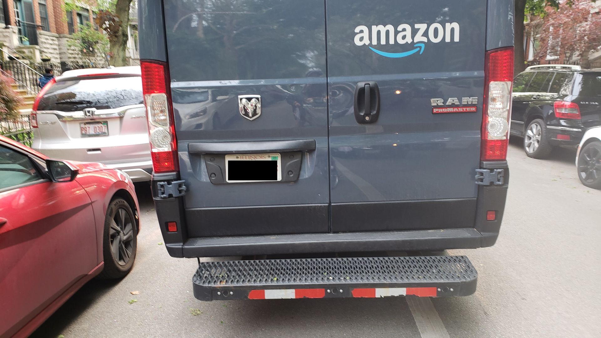 Rear view of a dark blue Amazon delivery van on a narrow urban street, flanked by a red and a silver car. The back doors show the white Amazon logo with a blue arrow; brick buildings and trees nearby.