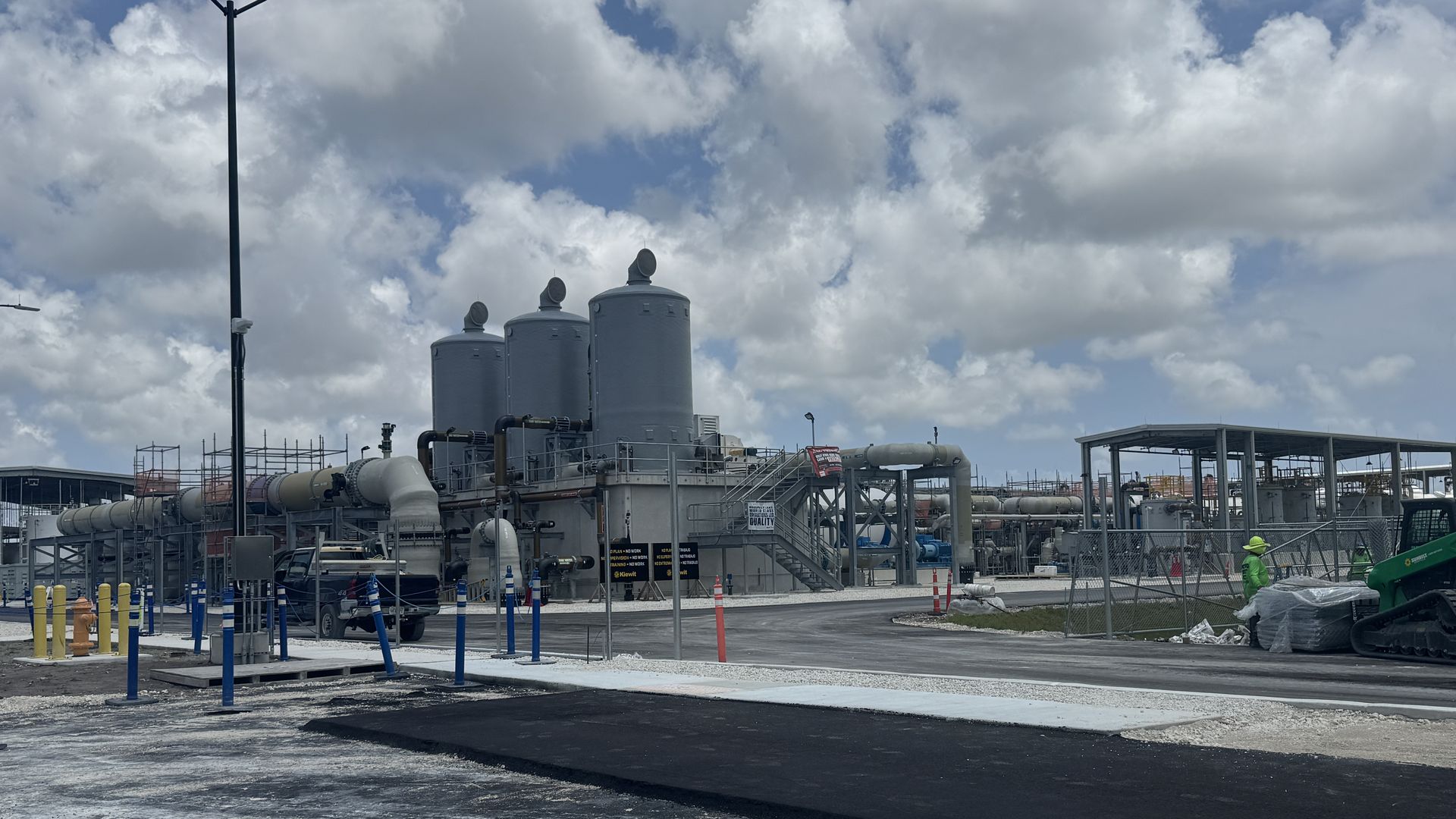 Water treatment with three tall grey silos, pipes, and metal scaffolding under a blue sky with clouds; workers visible bottom right.