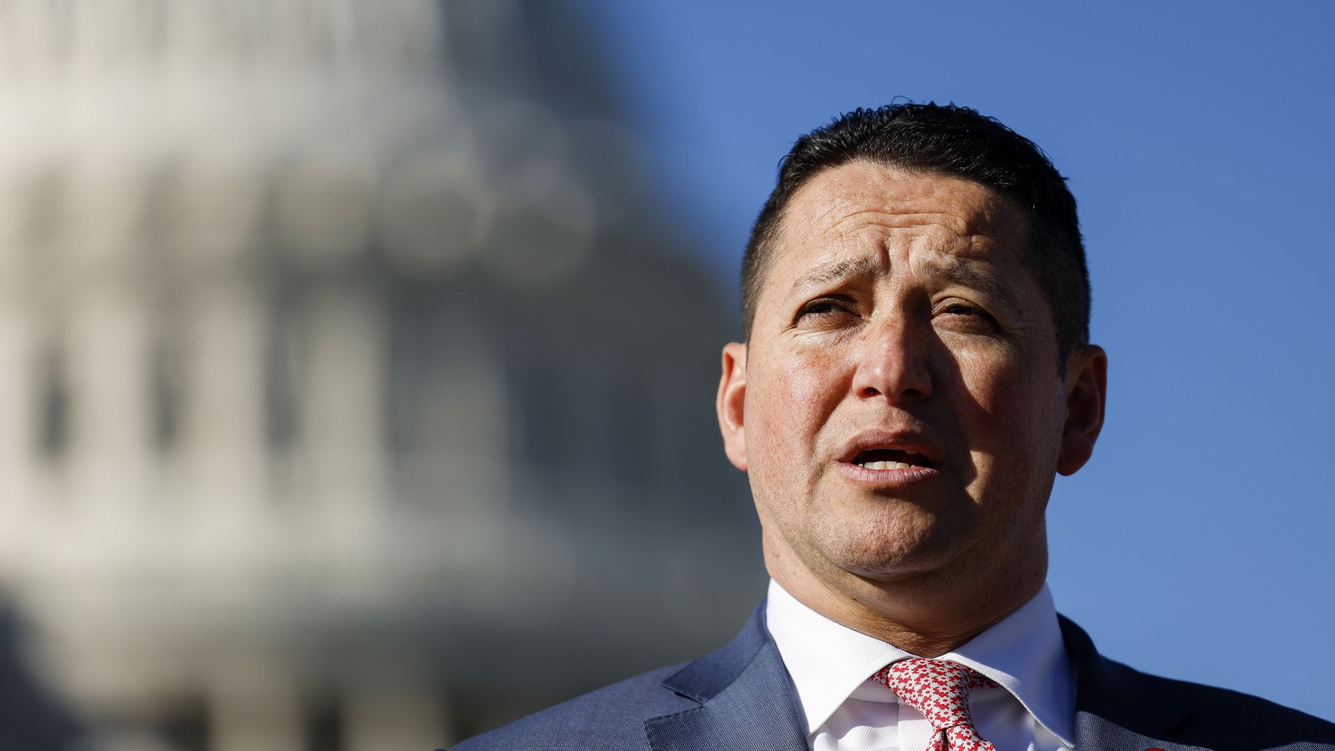 Close-up of a man in a blue suit and red patterned tie speaking outdoors with the U.S. Capitol building blurred in the background under a clear blue sky.