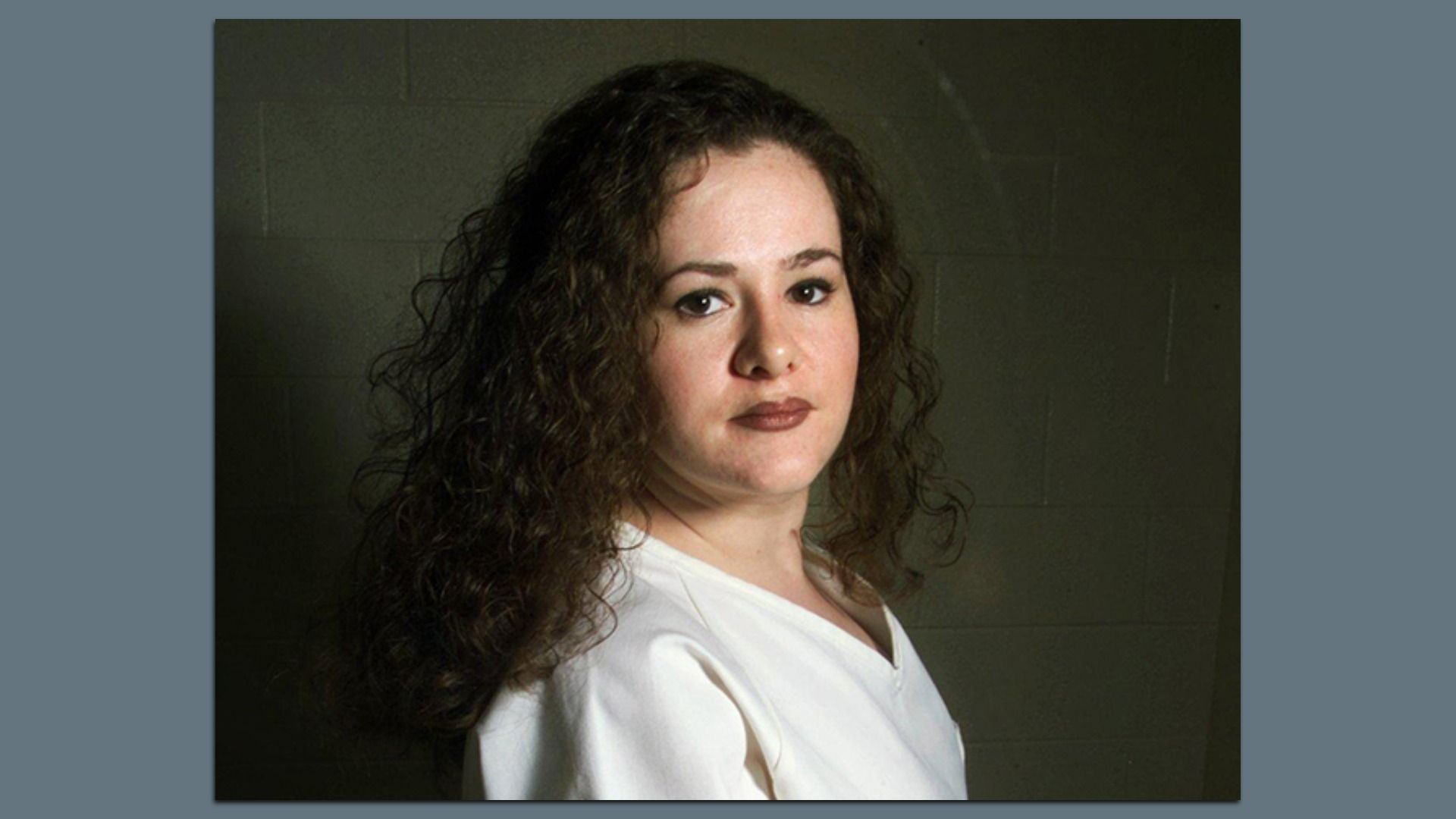 Head-and-shoulders portrait of a woman with long curly brown hair, wearing a white shirt, looking slightly to the right, against a plain gray wall.