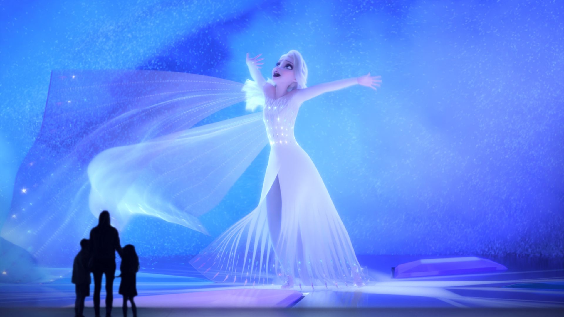A woman with two children stands in front of a massive screen showing Elsa from Frozen singing.