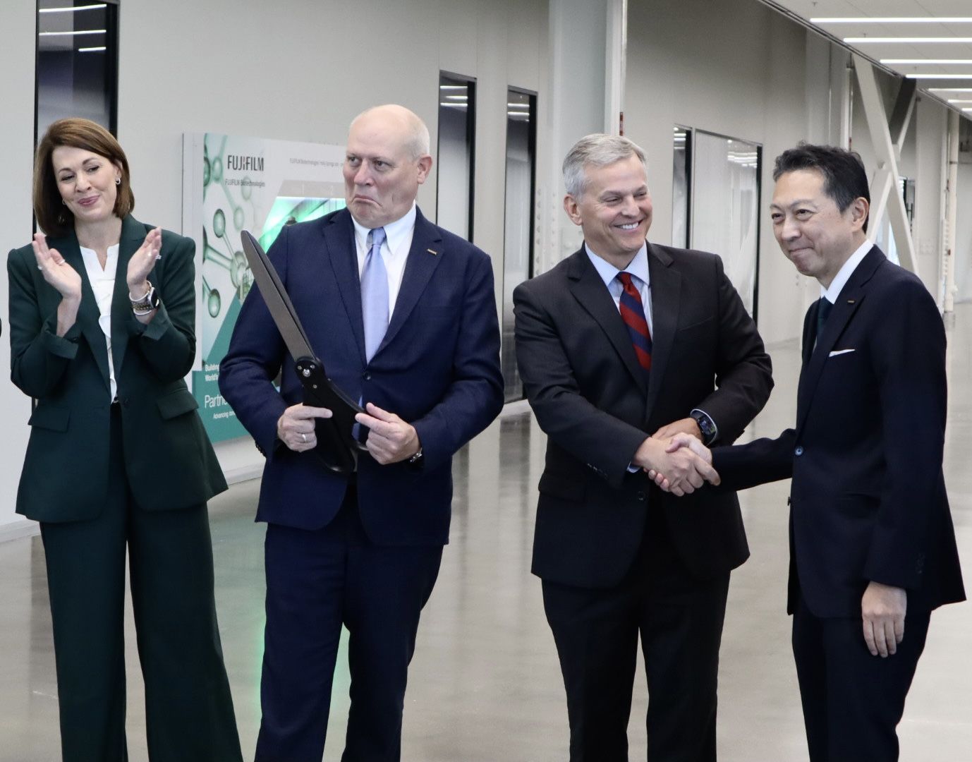 four people smile after a ribbon cutting indoors, one clapping, one holding large scissors, two shaking hands