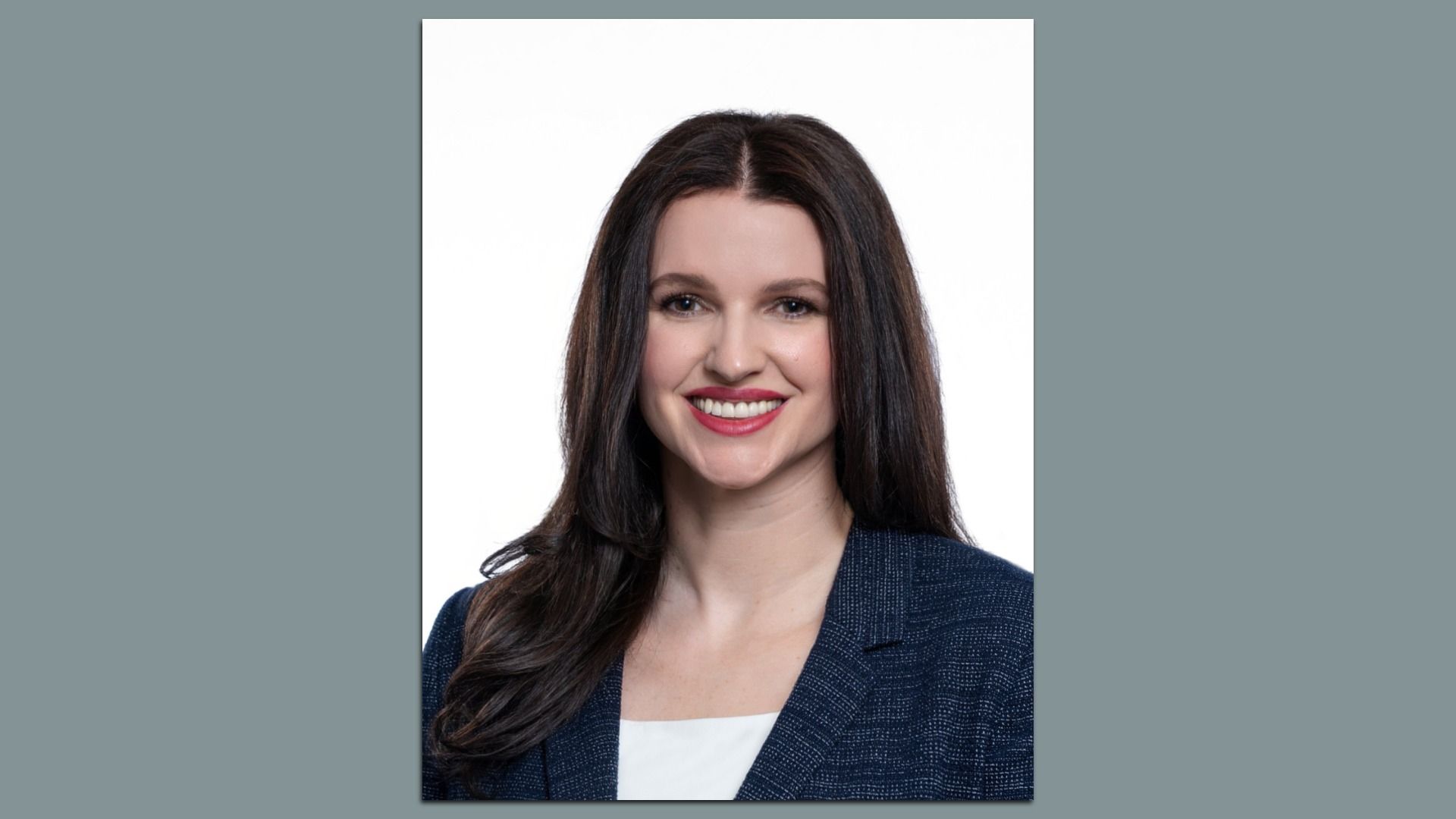 Portrait of a smiling woman with long dark hair, wearing a navy blazer over a white top, against a white background.
