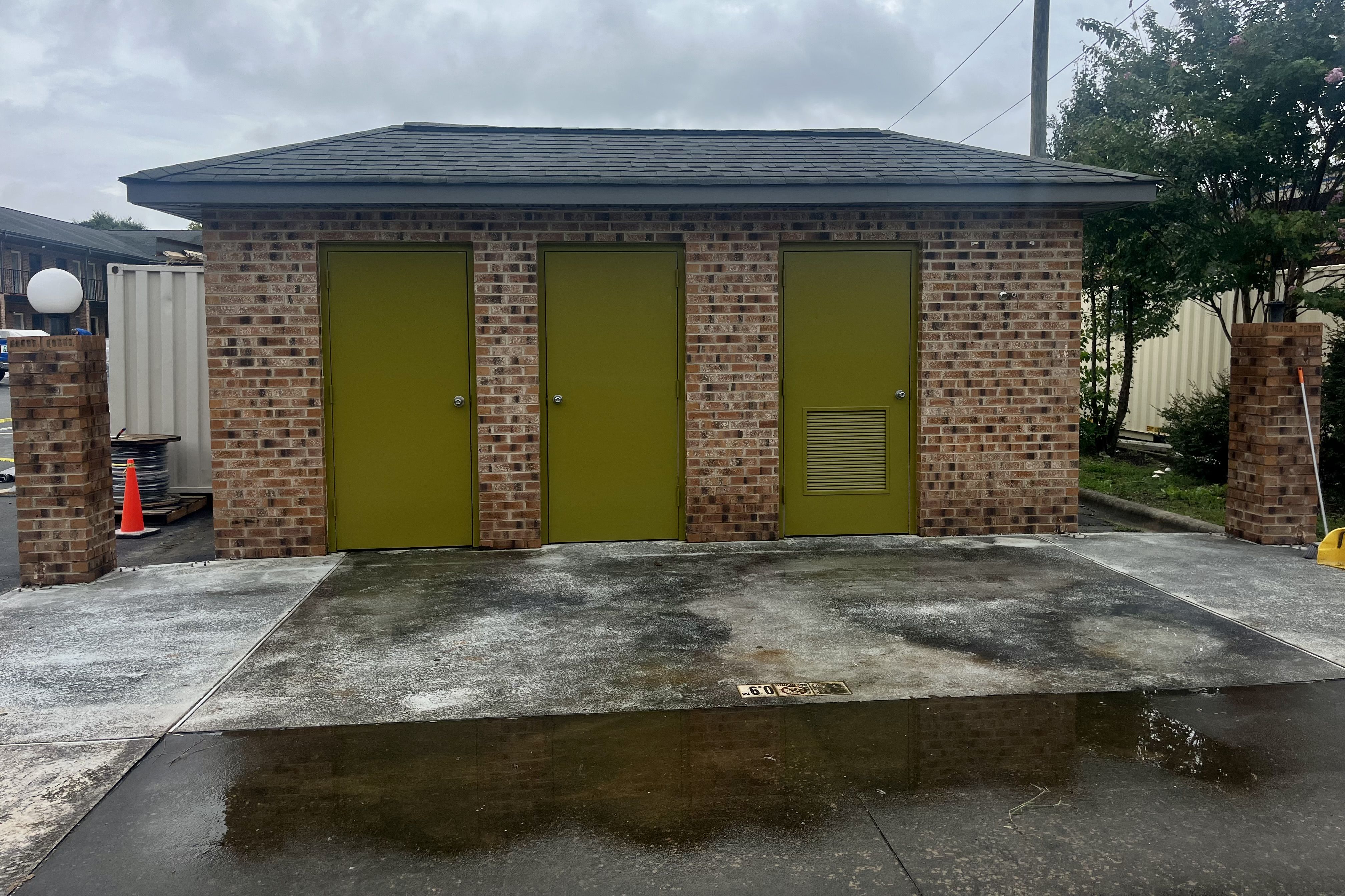 Brick building with three olive green doors, one with a vent, wet concrete ground in front reflecting part of the building, overcast sky, and trees on the right side.