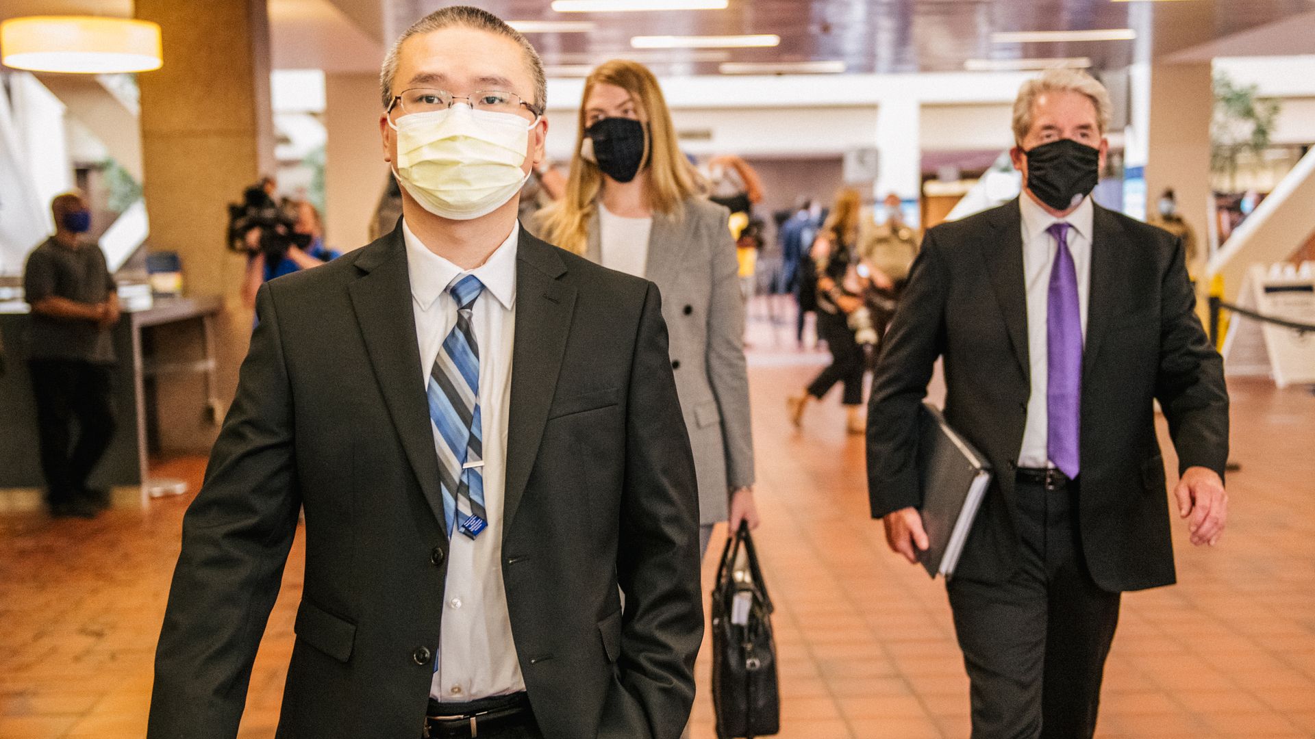 tou thao walks to court wearing a black suit and a blue striped tie and a mask and glasses. his lawyer and a woman, both in suits and a mask, walk behind him