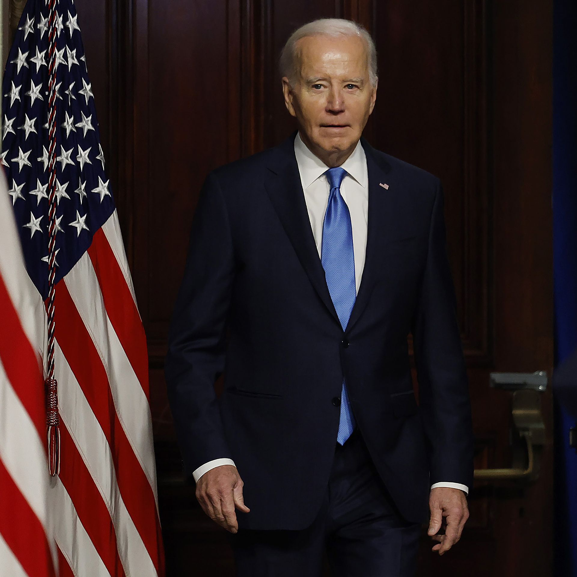 President Biden, in a dark suit, white shirt and blue tie, stands next to several American flags.