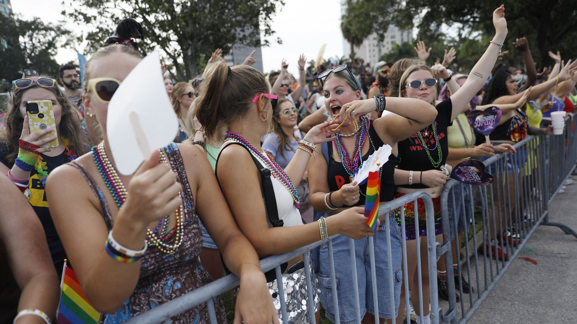 People with colorful beads and rainbow flags line a barricade fanning themselves.