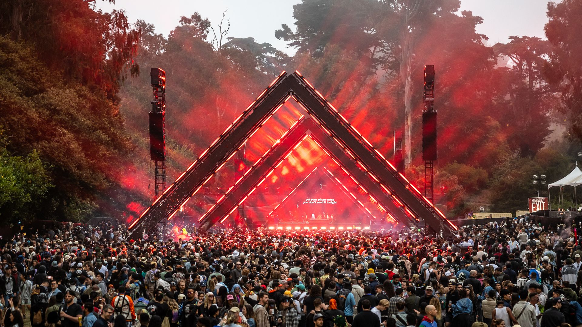 Large outdoor music festival crowd in front of a stage with red triangular light structures and red lighting effects, surrounded by tall trees under a gray sky.