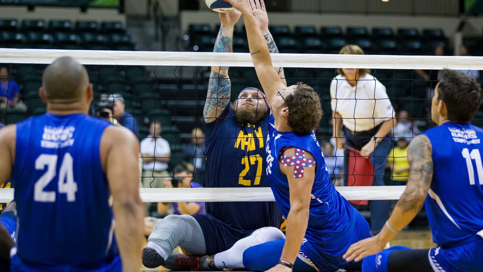 Wounded military members and veterans play volleyball while sitting on the ground, with their hands in the air.