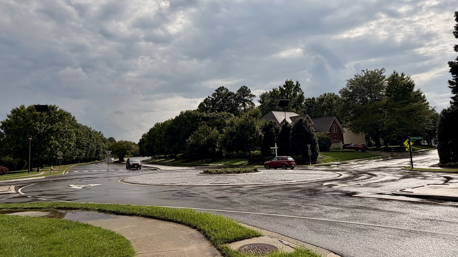 Overcast sky above a wet suburban roundabout with a circular central island, red and dark cars, green trees lining the road, and brick houses in the background.