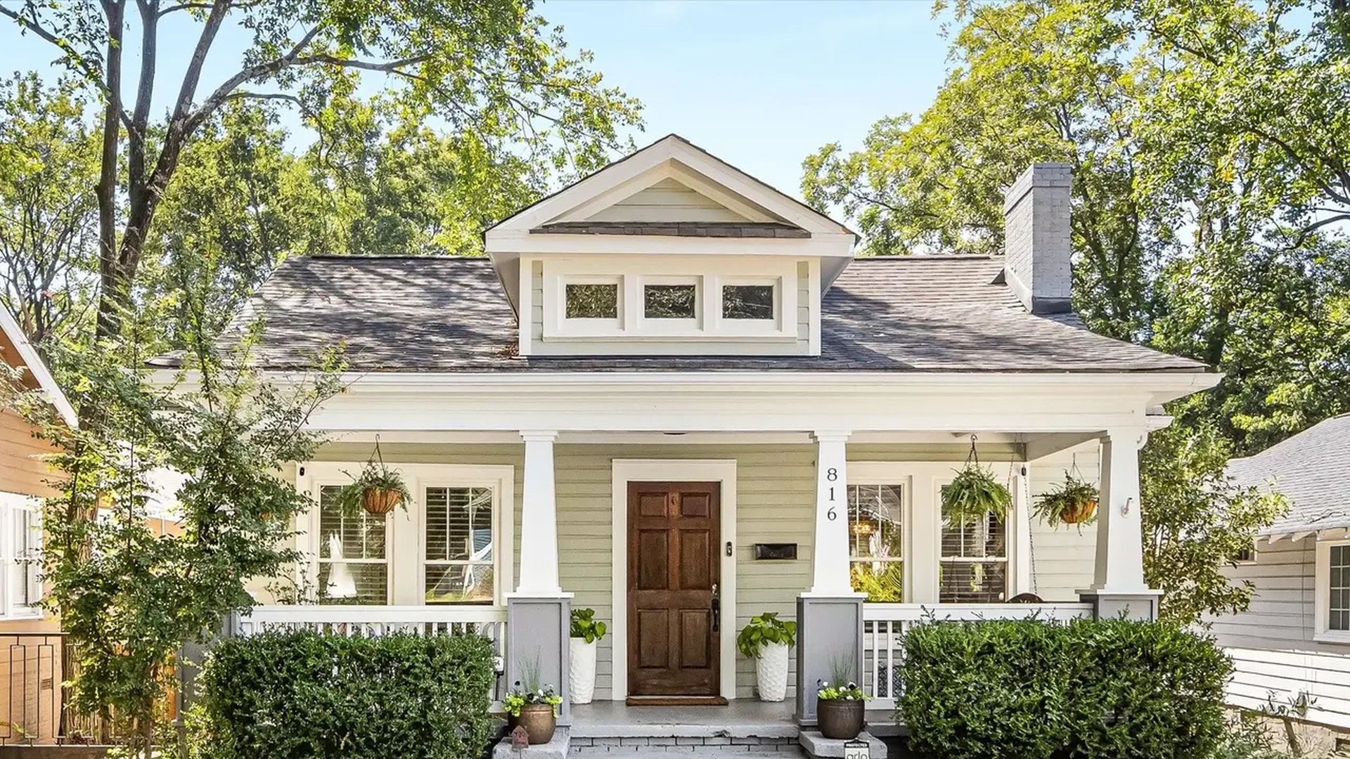 Light green bungalow house with white trim, brown front door, hanging plants on porch, bushes along walkway, and address number 816 on column.