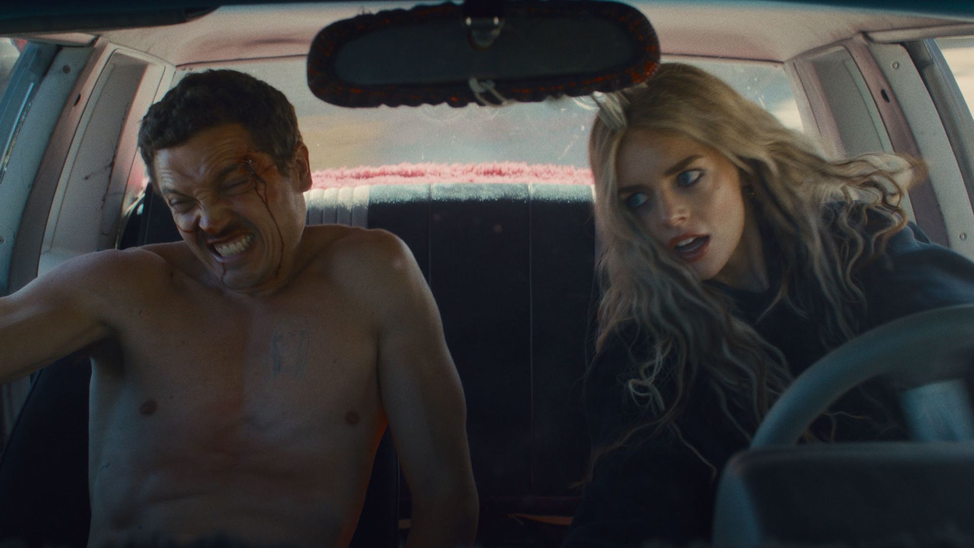 Inside a moving car, a shirtless man with a bleeding head injury winces in pain while a woman with long blonde hair drives, looking concerned and focused on the road ahead.