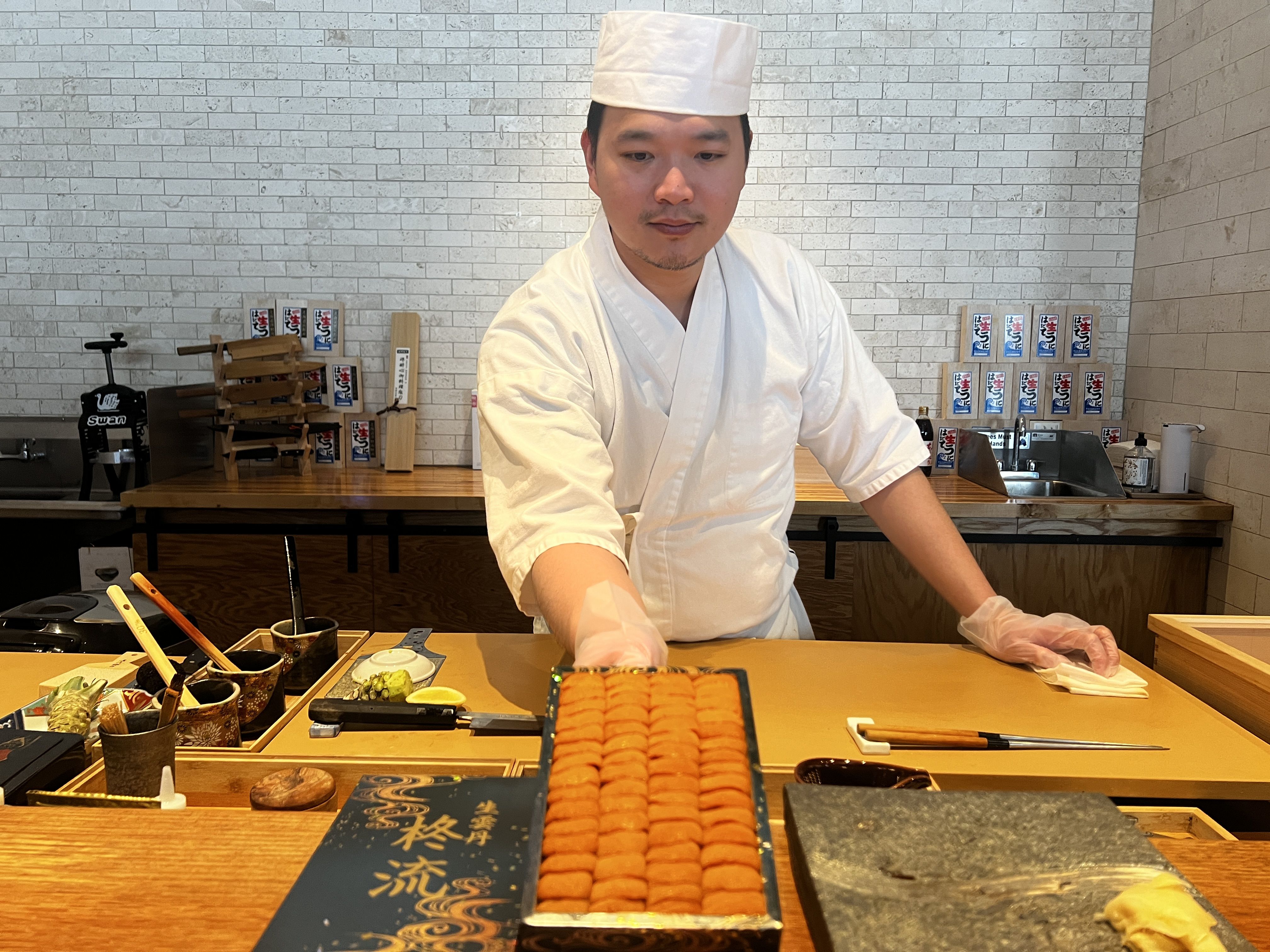 A sushi chef wearing a white uniform and cap is standing behind a counter. He is presenting a tray filled with neatly arranged pieces of uni (sea urchin). The counter has various items such as small bowls, utensils, and garnishes, including wasabi and lemon slices. 