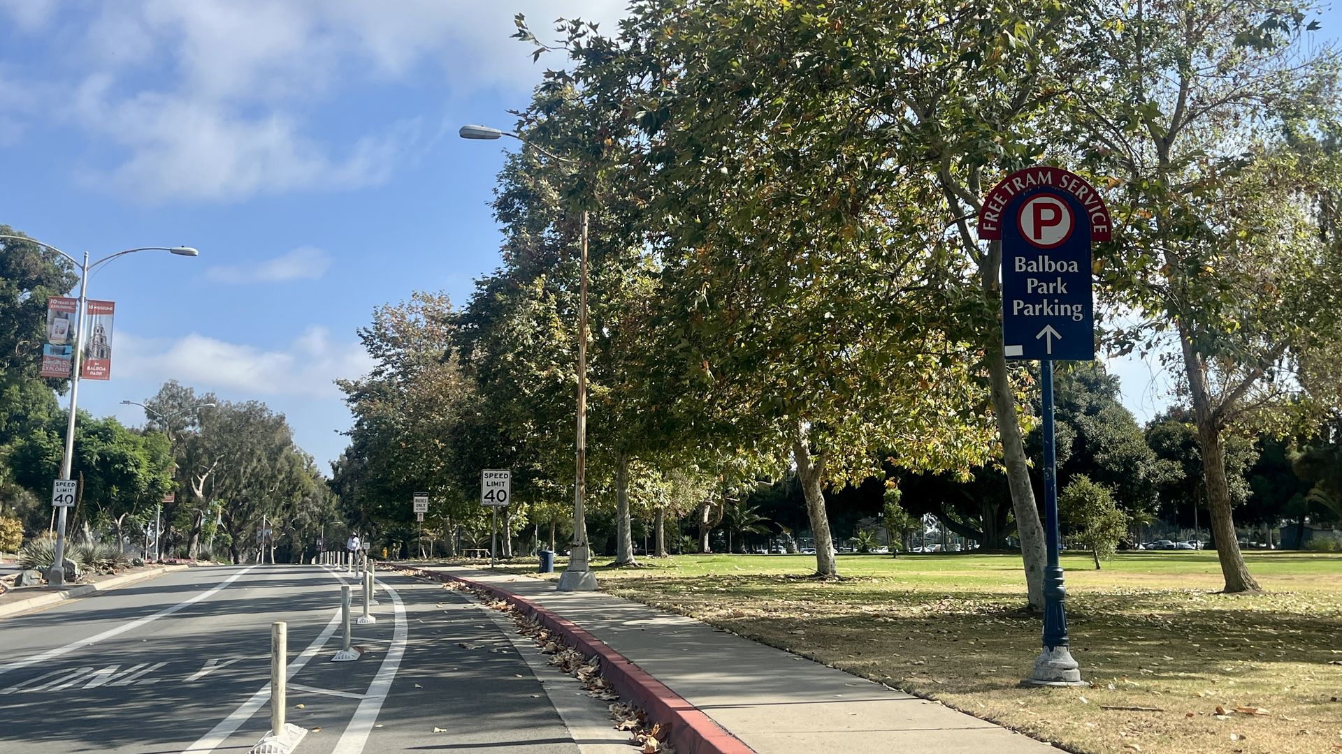 Sunny street scene with trees and blue sky, a sign reading "Free Tram Service Balboa Park Parking" along a sidewalk next to a road.