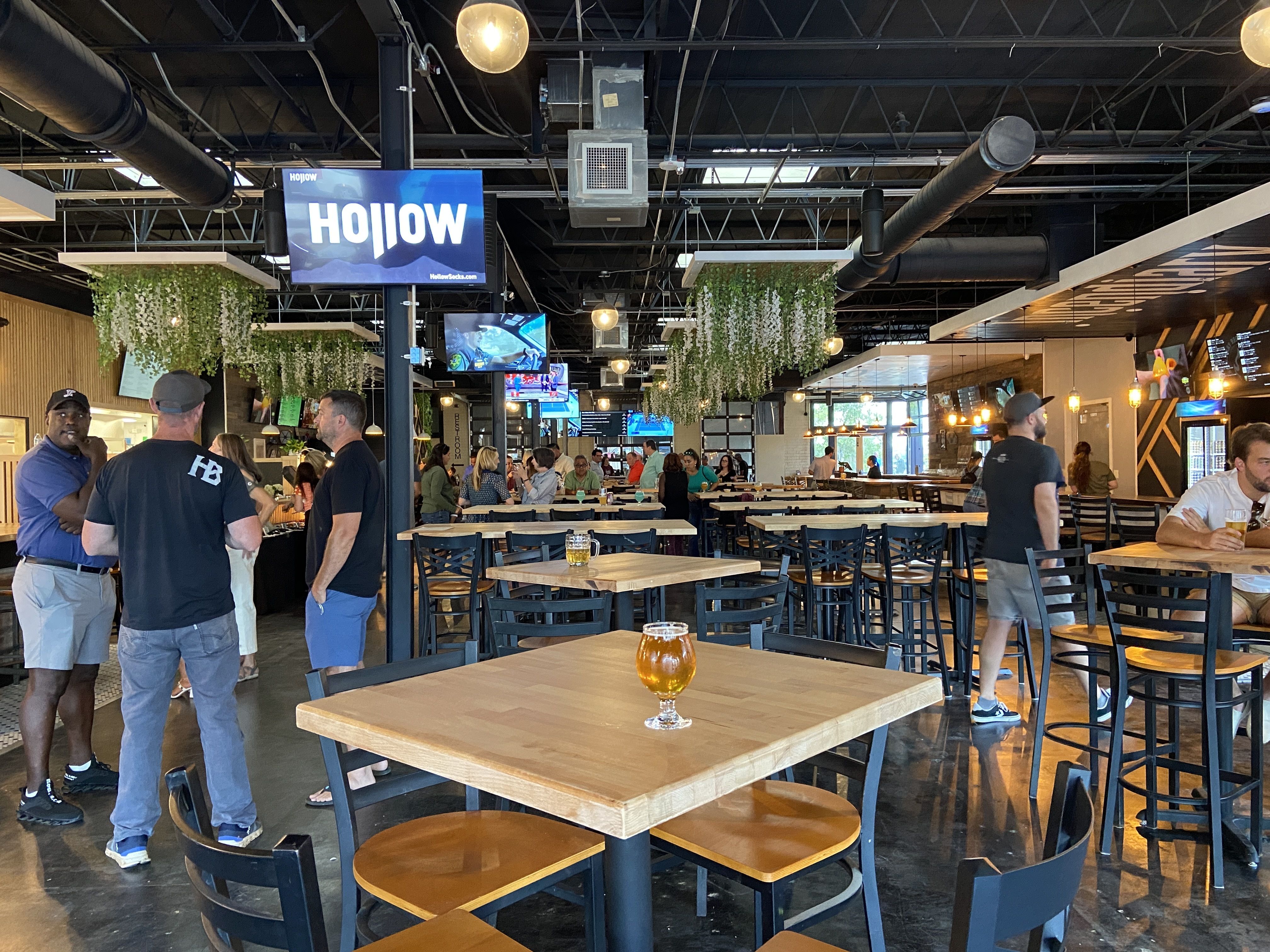 Interior of a modern brewery taproom with wooden tables and black chairs, hanging greenery, TVs showing sports, and people talking and enjoying drinks in a casual setting.