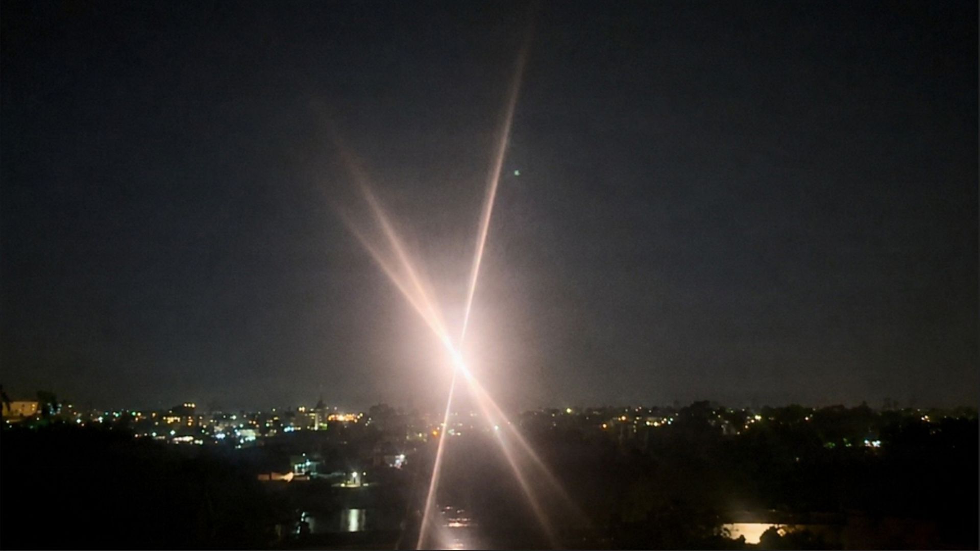 Night cityscape at night. A bright flash at the center sends intersecting light trails upward, illuminating the sky.
