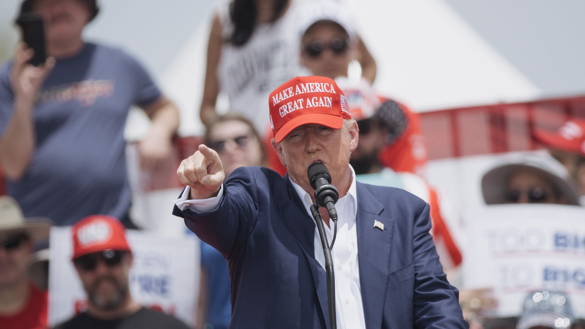 Donald Trump, wearing a red "Make America Great Again" hat and a blue suit with no tie, speaking into a microphone and pointing at a crowd at a rally.