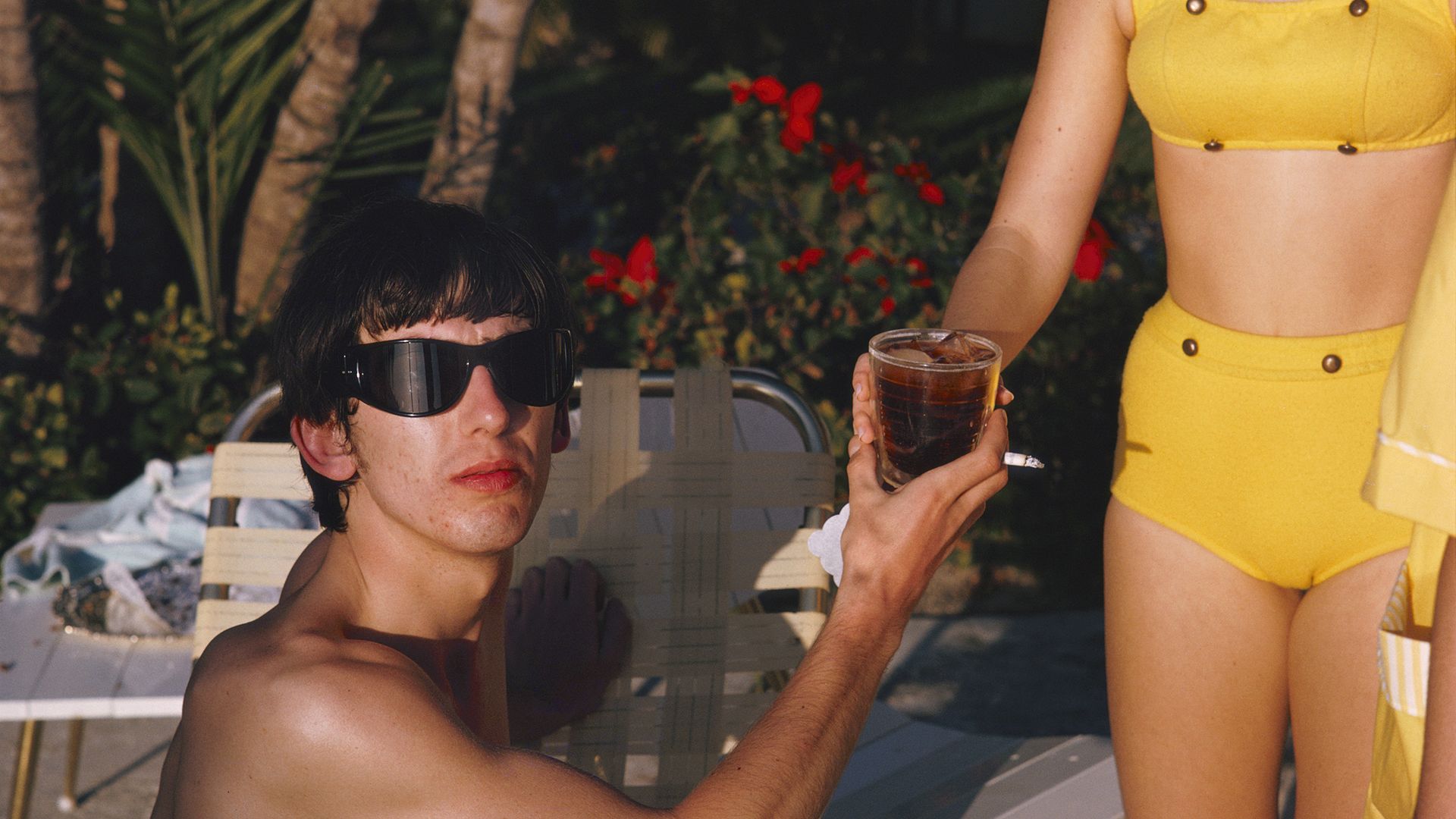 A photo of a man wearing sunglasses facing toward a camera and receiving a beverage from a person wearing a yellow bathing suit.