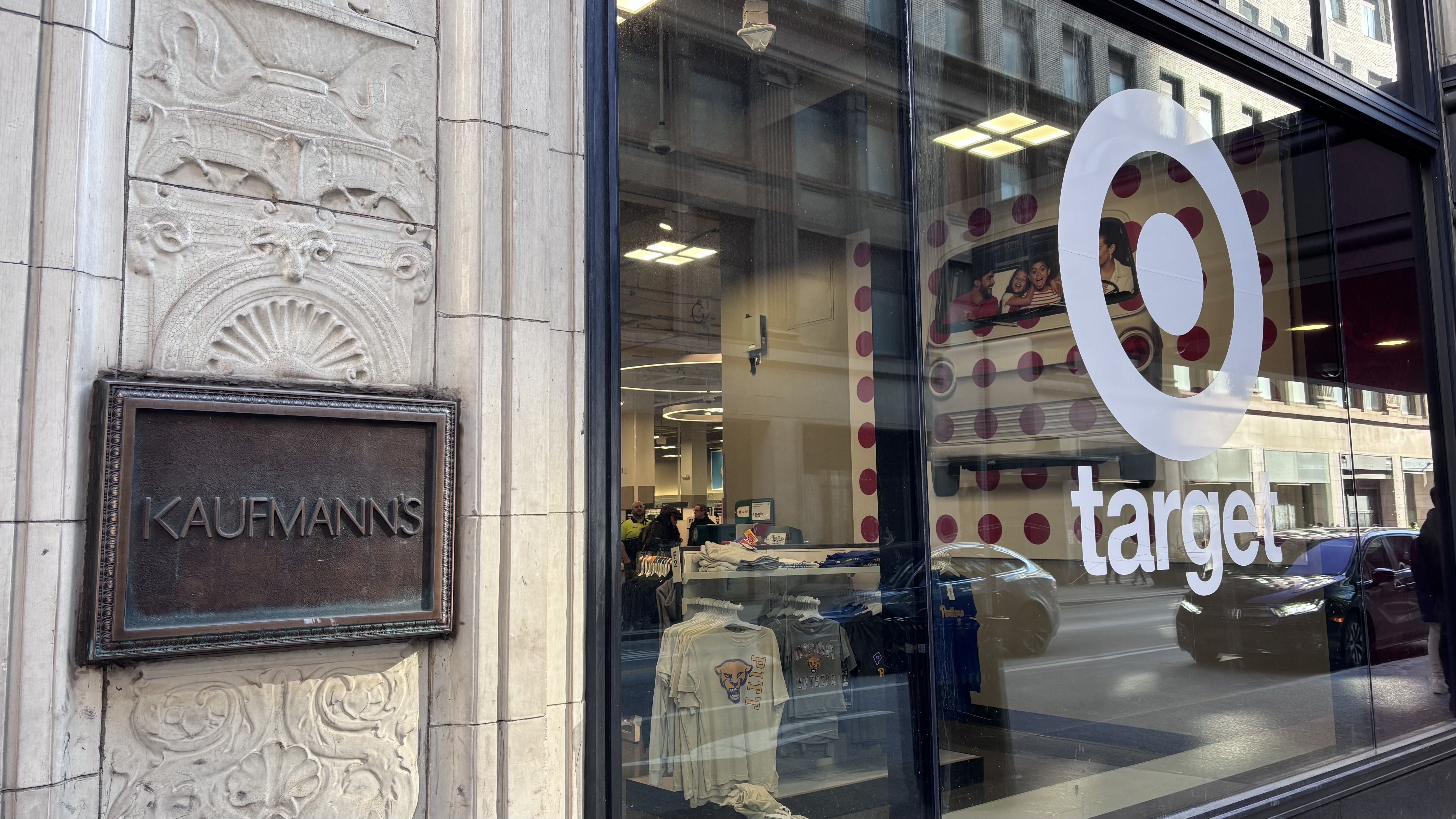 Street view of a Target store window with large white Target logo, reflections of cars on street, and a bronze plaque reading "KAUFMANN'S" mounted on ornate stone wall beside the window.