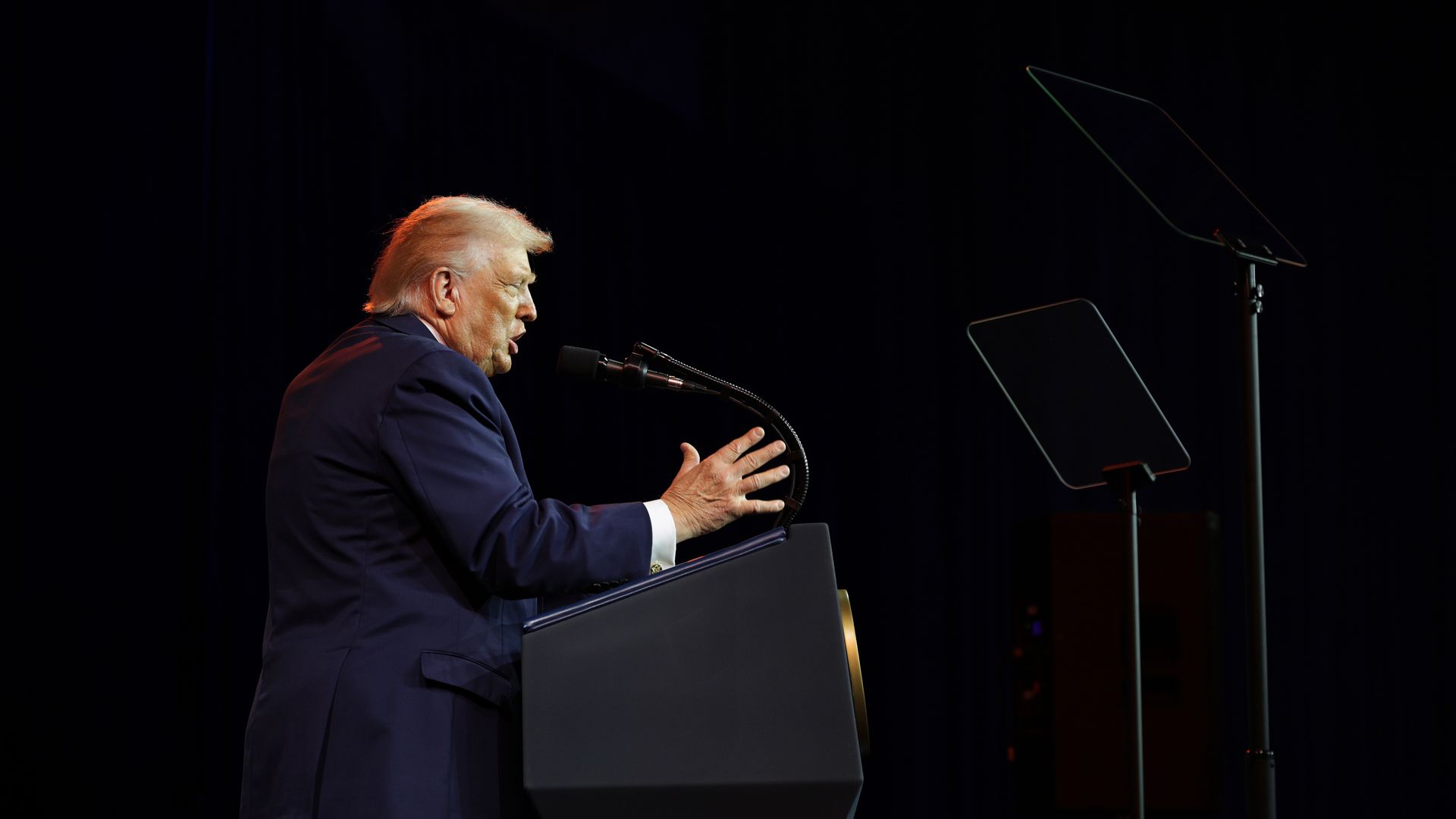 Trump standing at a podium speaking into a microphone with one hand raised, wearing a navy suit jacket and white shirt
