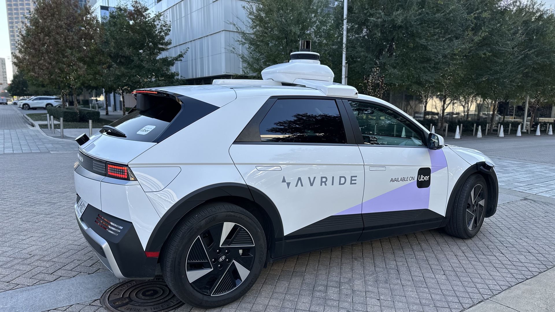 White and purple autonomous Avride vehicle with Uber logo parked on tiled pavement near urban buildings and trees in Dallas.