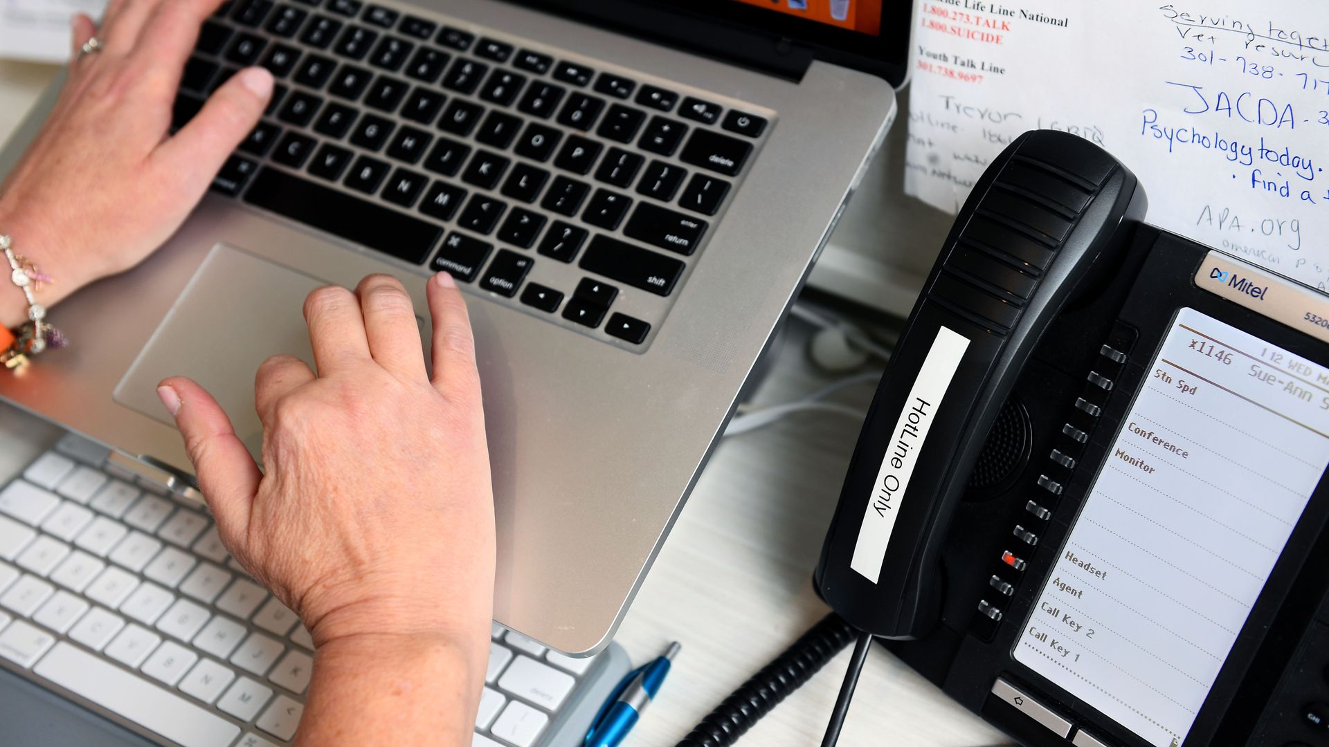 two hands touch the keyboard of a laptop while a black landline phone sits next to it