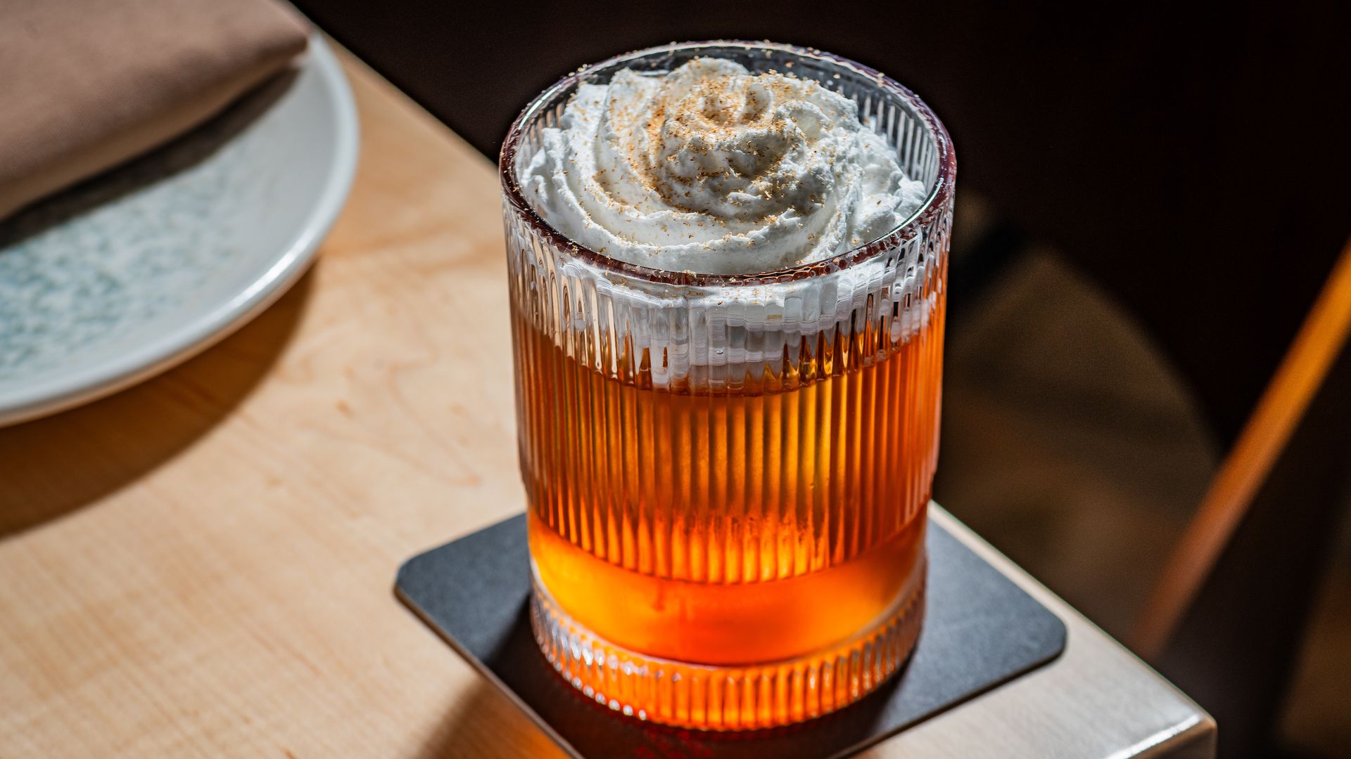 Amber, ribbed glass filled with a drink topped with whipped cream and a light dusting of cinnamon, on a metal coaster beside a wooden table with a blurred plate in the background.