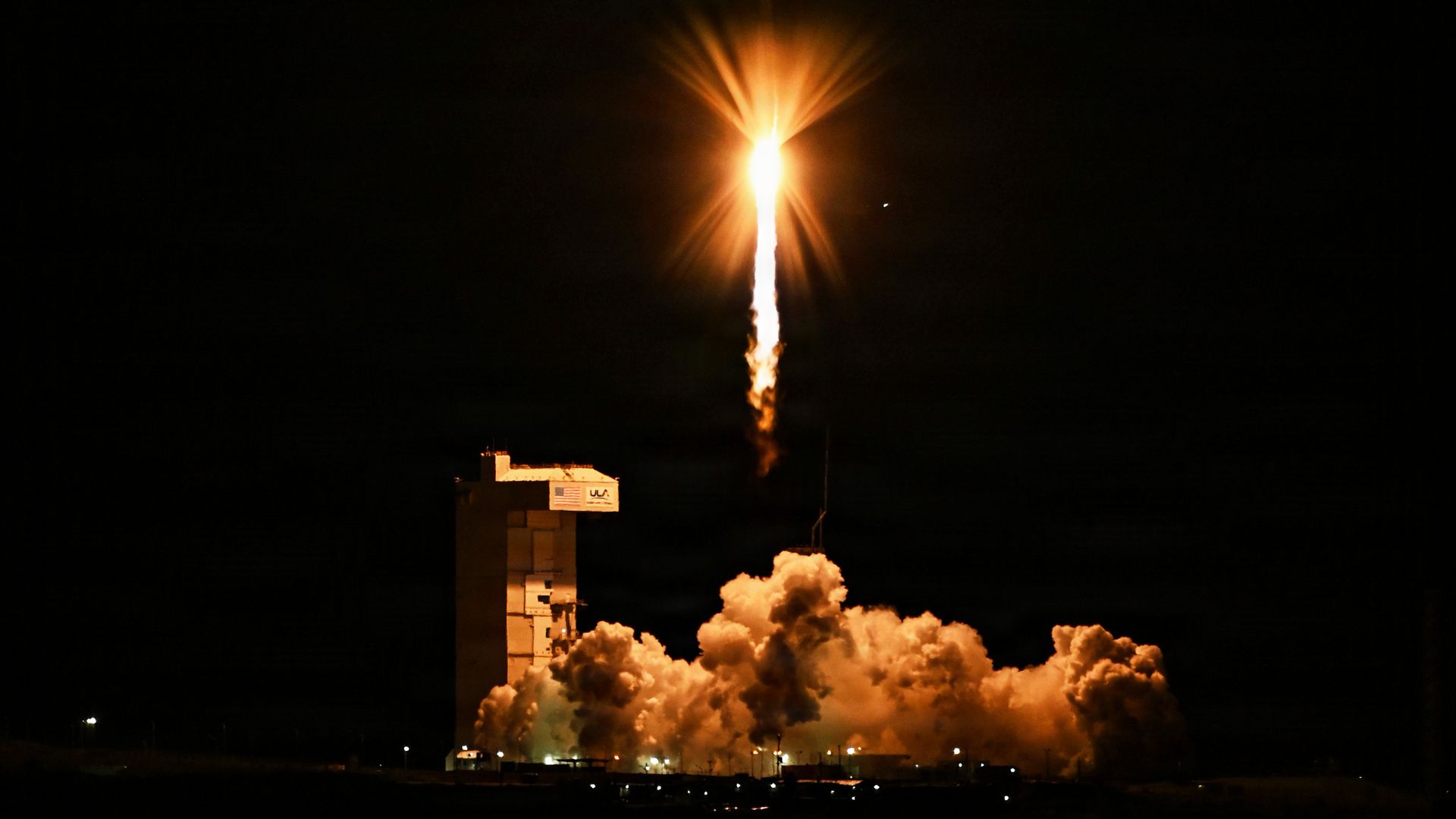 A bright rocket launches at night, orange flame and radiant exhaust plume shooting upward, with billowing smoke near the pad and a dark sky; a launch complex silhouette at the bottom.