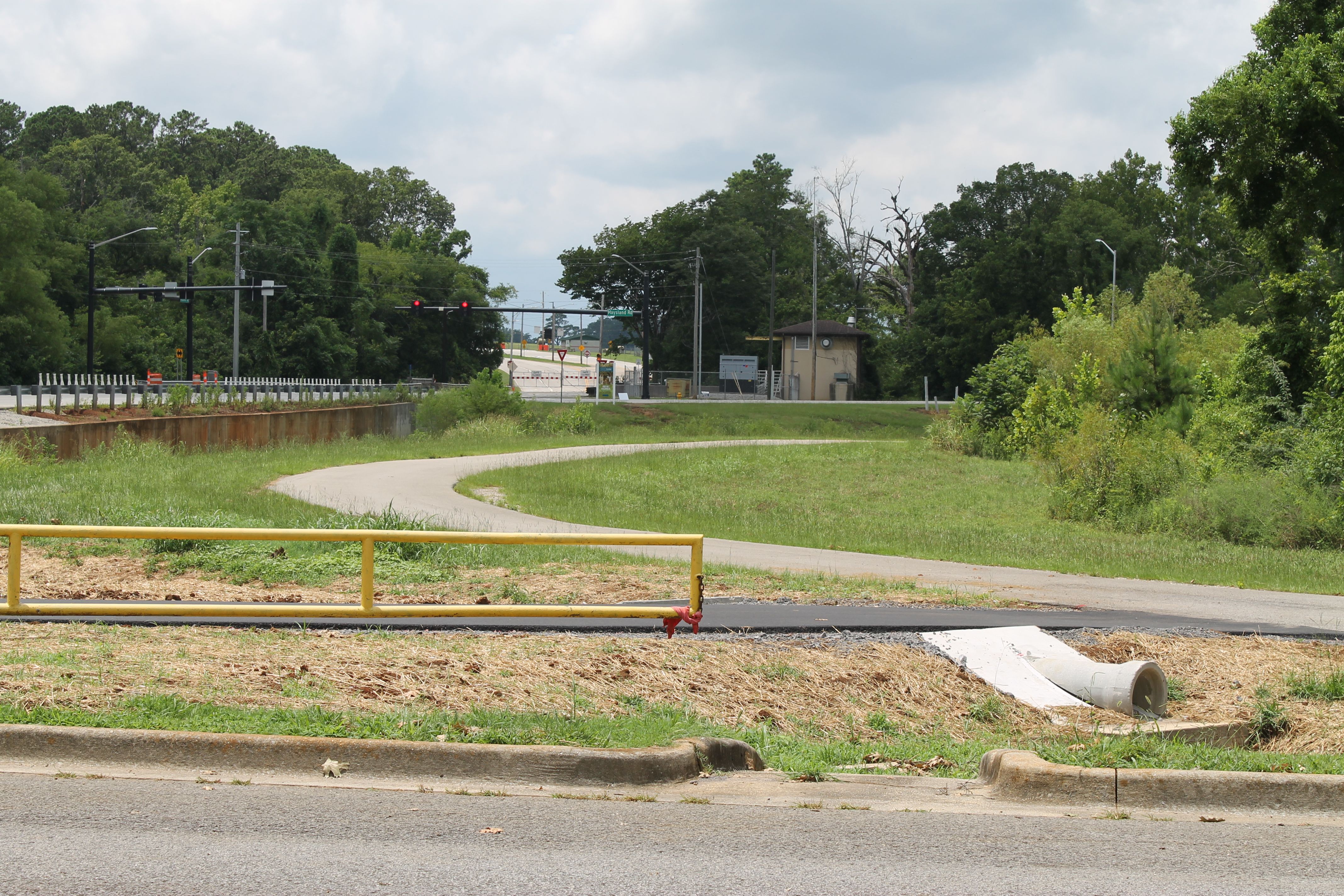 Wide roadside scene with a yellow guard rail in the foreground, a curved path through grass, a construction area with red traffic lights, and a small beige utility shed among trees.