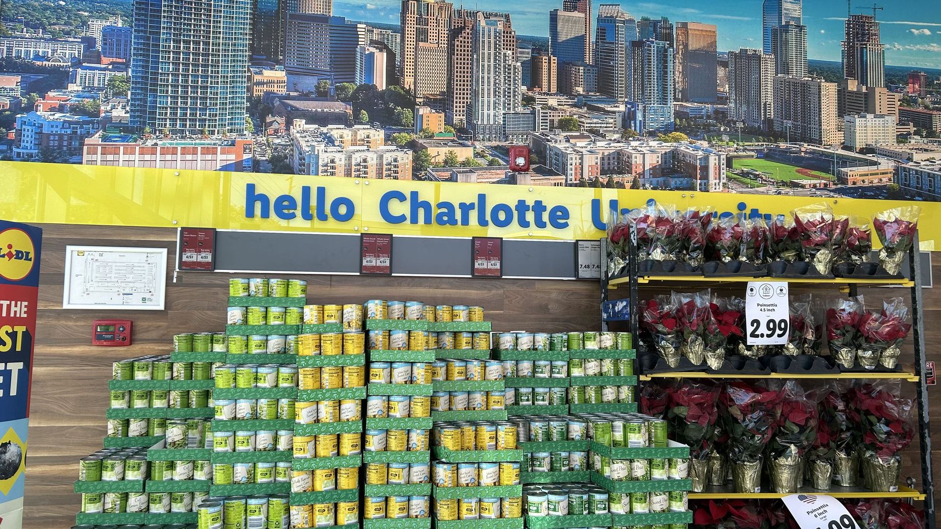 Display of canned goods stacked under a large Charlotte cityscape poster with the text "hello Charlotte University". To the right, potted red poinsettia plants are for sale priced at $2.99 and $4.99.