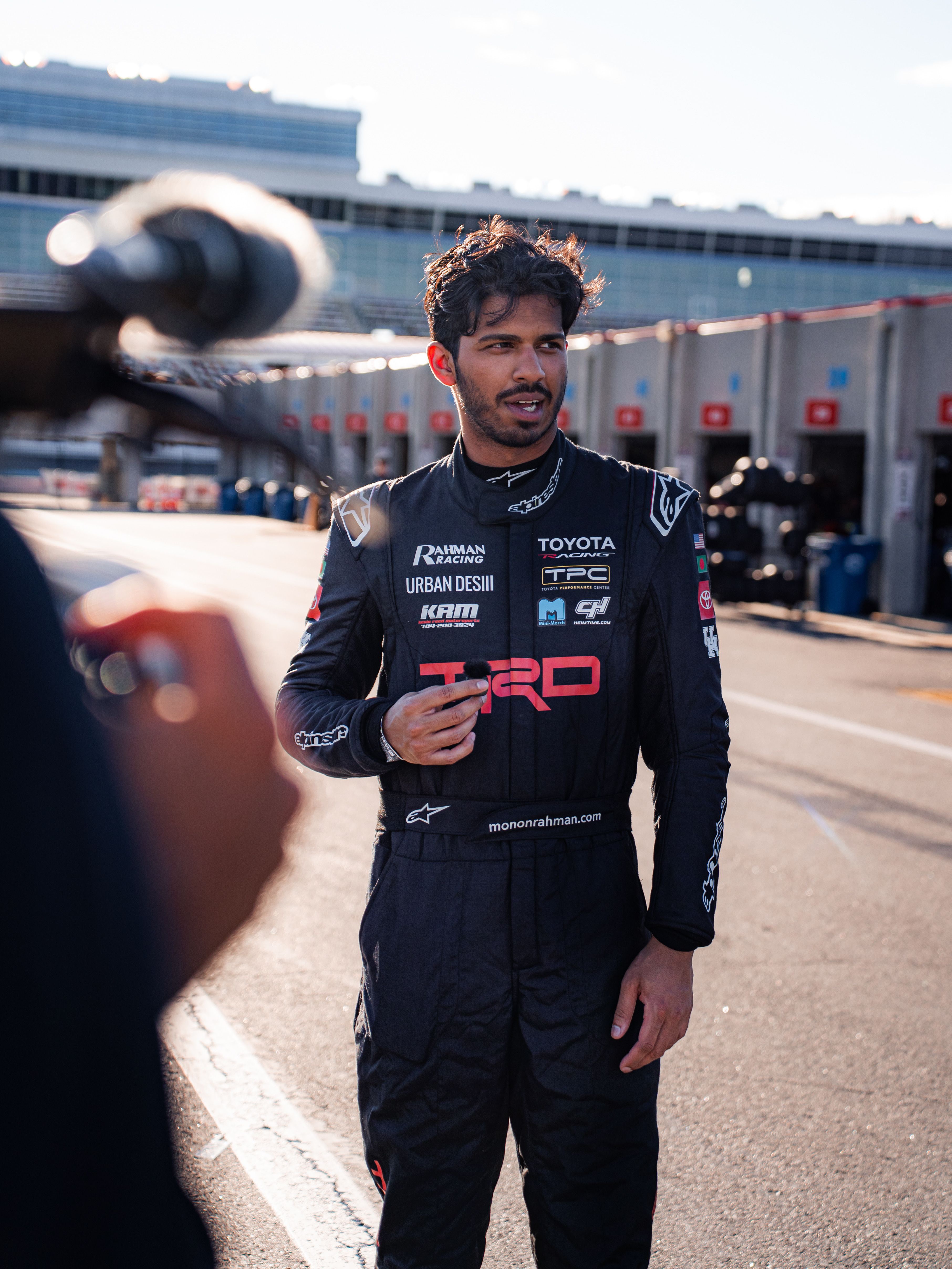 Young man in a black racing suit with sponsor logos stands in a sunlit pit lane, speaking into a small microphone; dark hair and beard, with blurred garages behind.