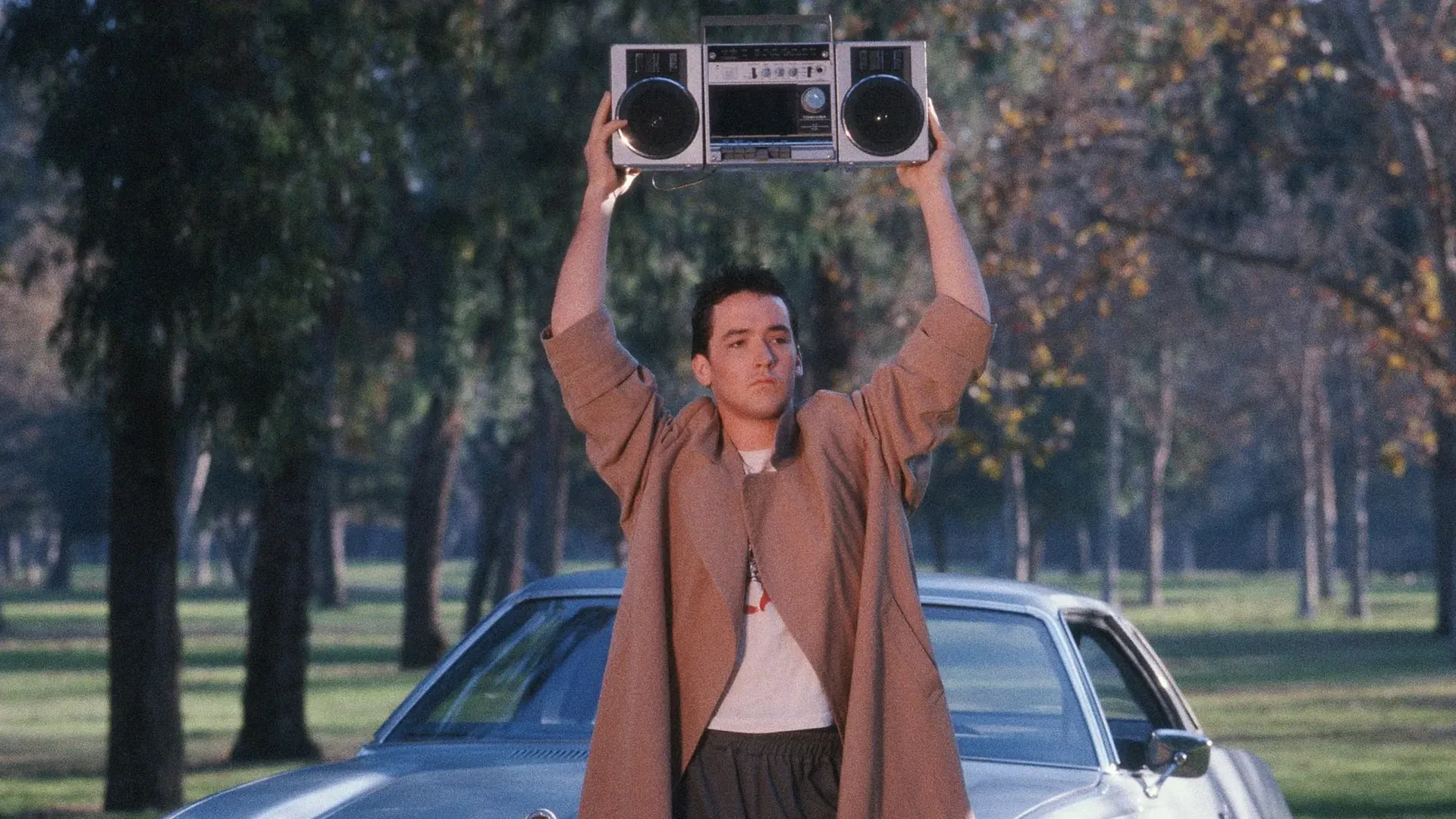 A man in a long brown coat stands in a park holding a boombox above his head with a silver car behind him, trees and grass in the background during the day.