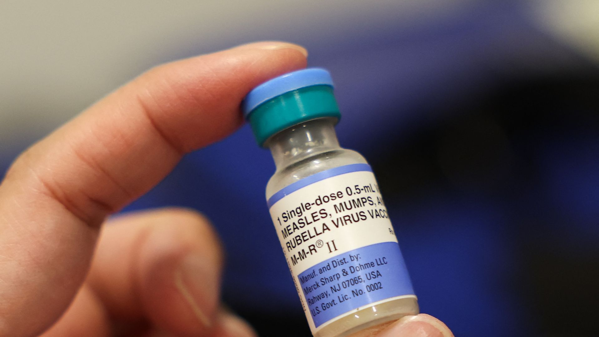 A vial of the Measles, Mumps and Rubella vaccination for children with a light purple and blue cap is held between a person's thumb and index finger