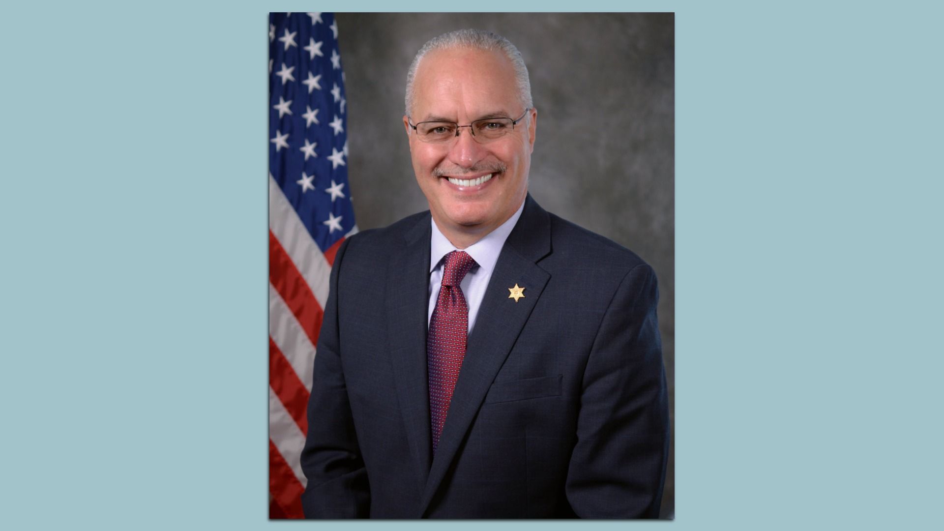 Sheriff Hall in a navy suit, light shirt, and red tie with a gold star badge on his lapel, posed in front of the U.S. flag and a gray backdrop.