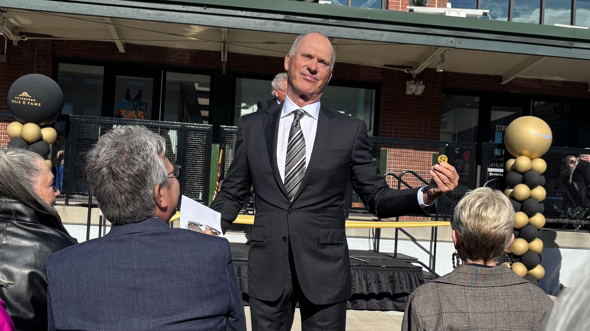 Man in black suit and striped tie holding a small yellow token, standing outdoors addressing an audience with black and gold balloon columns labeled "Pittsburgh Walk of Fame" in the background.