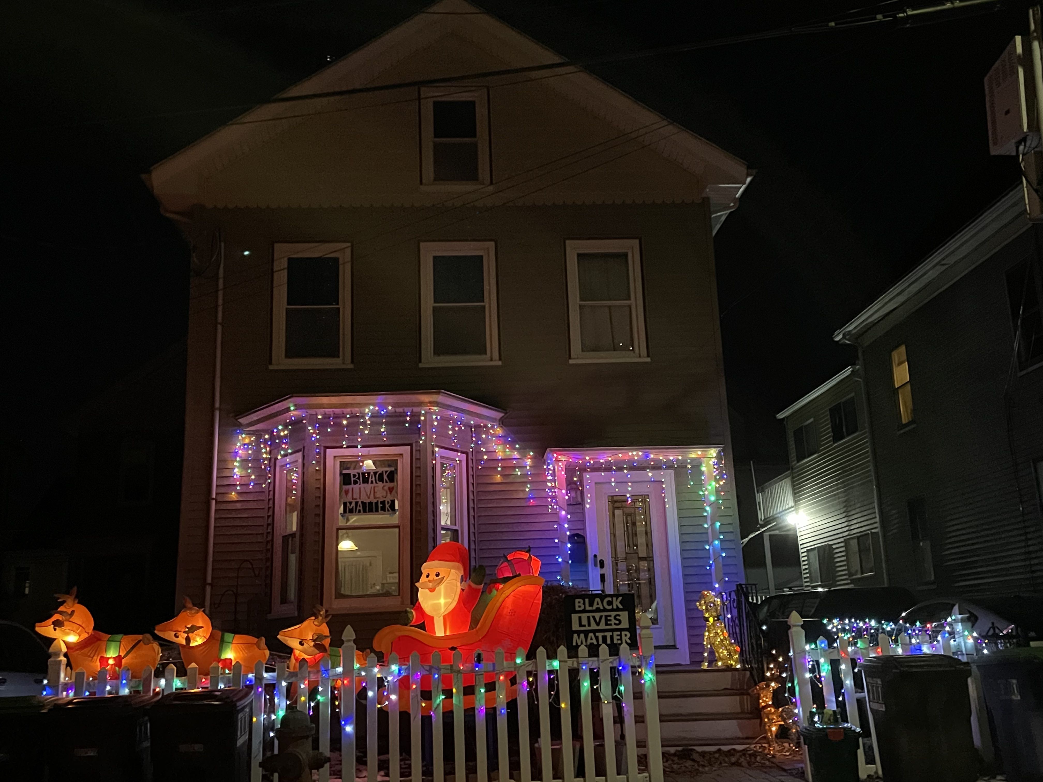A large inflatable Santa on a sleigh behind three reindeer stand on the front lawn of a lit-up house with Black Lives Matter signs.