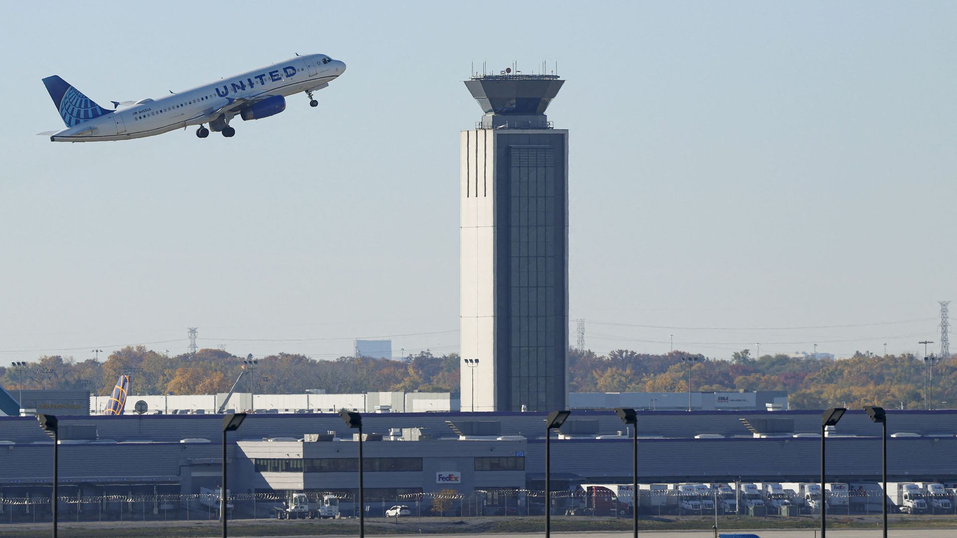 United Airlines jet climbs off a runway beside a tall airport control tower; white fuselage with blue UNITED lettering and blue tail, with terminal buildings and a clear sky in the background.