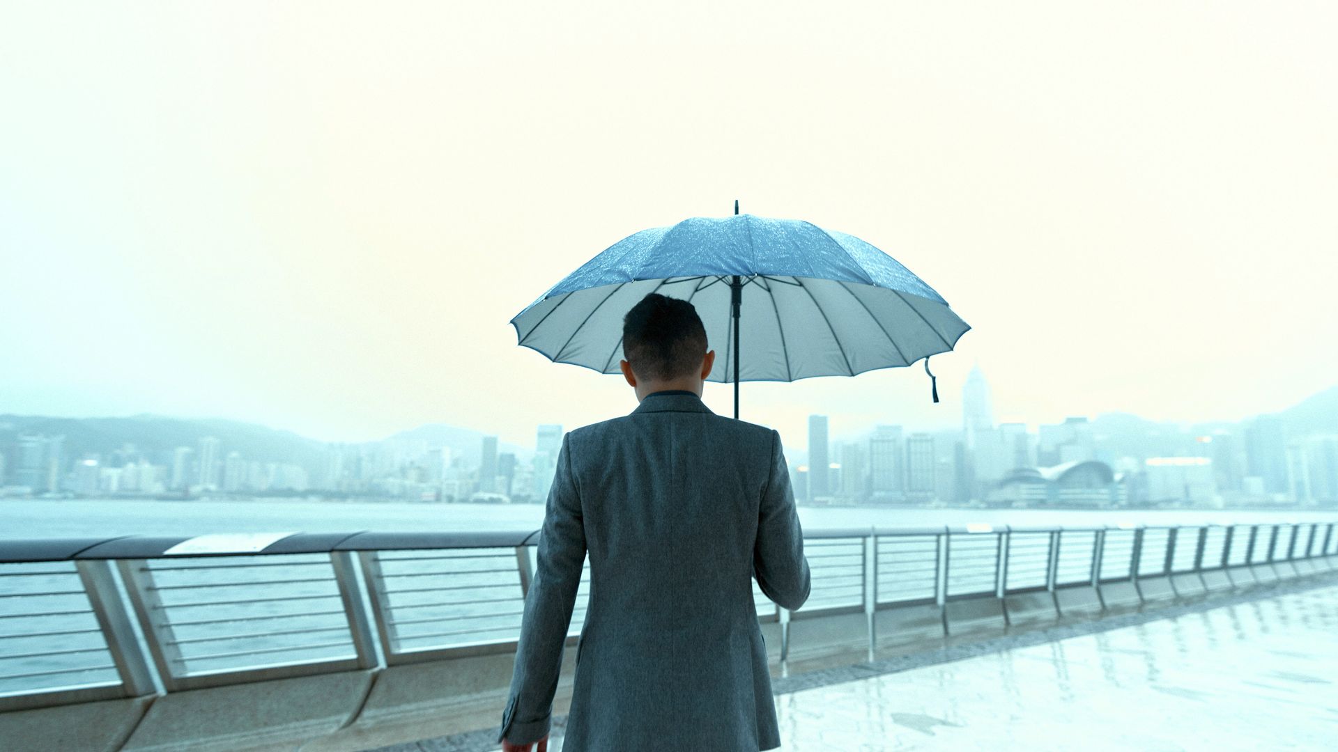 An image of a man from behind, outside on a bridge, holding an umbrella, in front of a cityscape.
