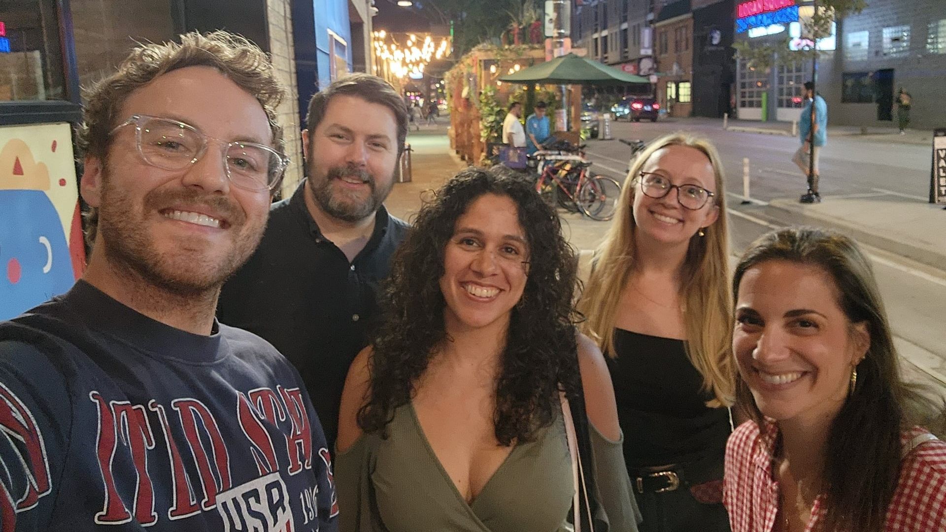 Group of five friends smiling for a selfie on a lively city street at night. Warm street lights, string lights, a green umbrella cafe, bikes, and colorful storefronts in the background.