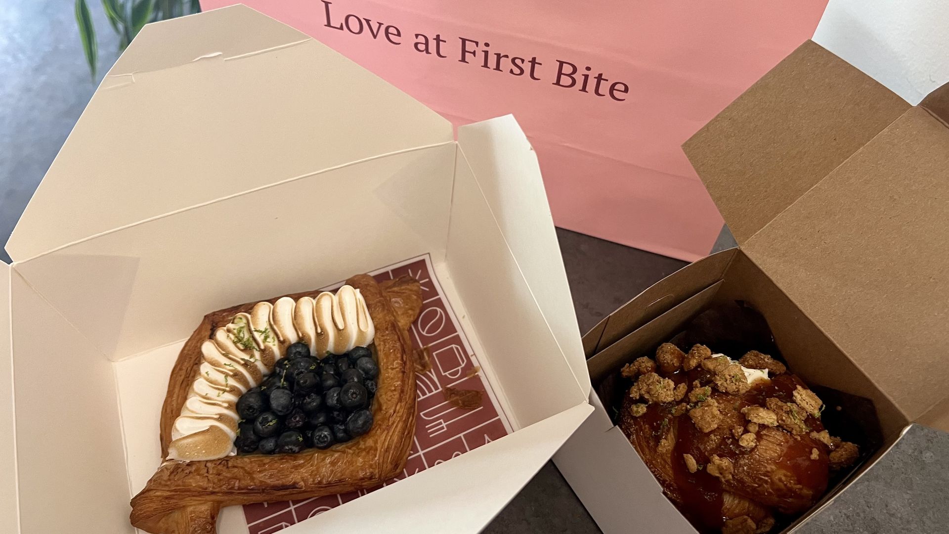 Two pastries in boxes on a table: one with blueberries and cream on a golden puff pastry, the other topped with crumble and glaze, in front of a pink bag saying "Love at First Bite".