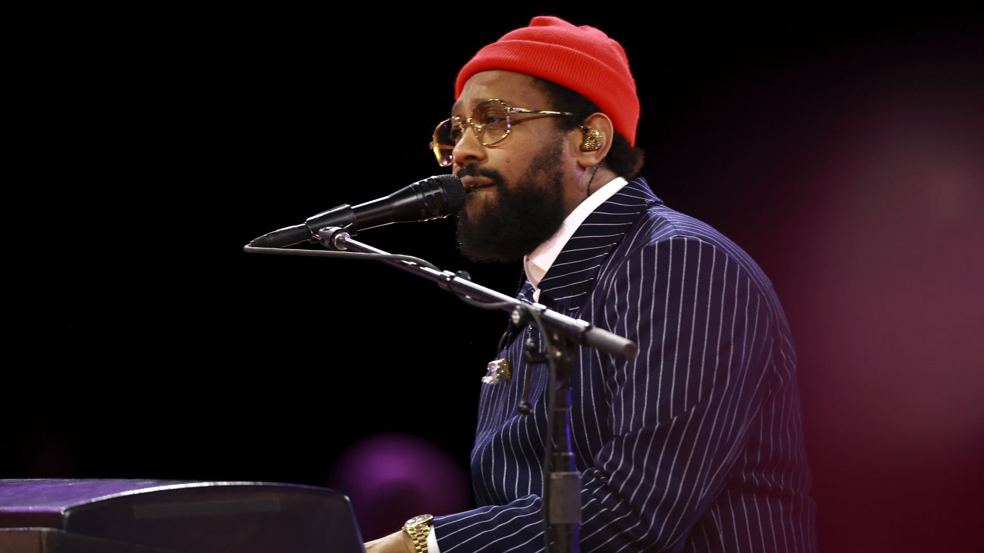 Photo shows PJ Morton singing into a microphone and playing a piano. He wears a pinstripe jacket, glasses, gold watch and a red beanie.