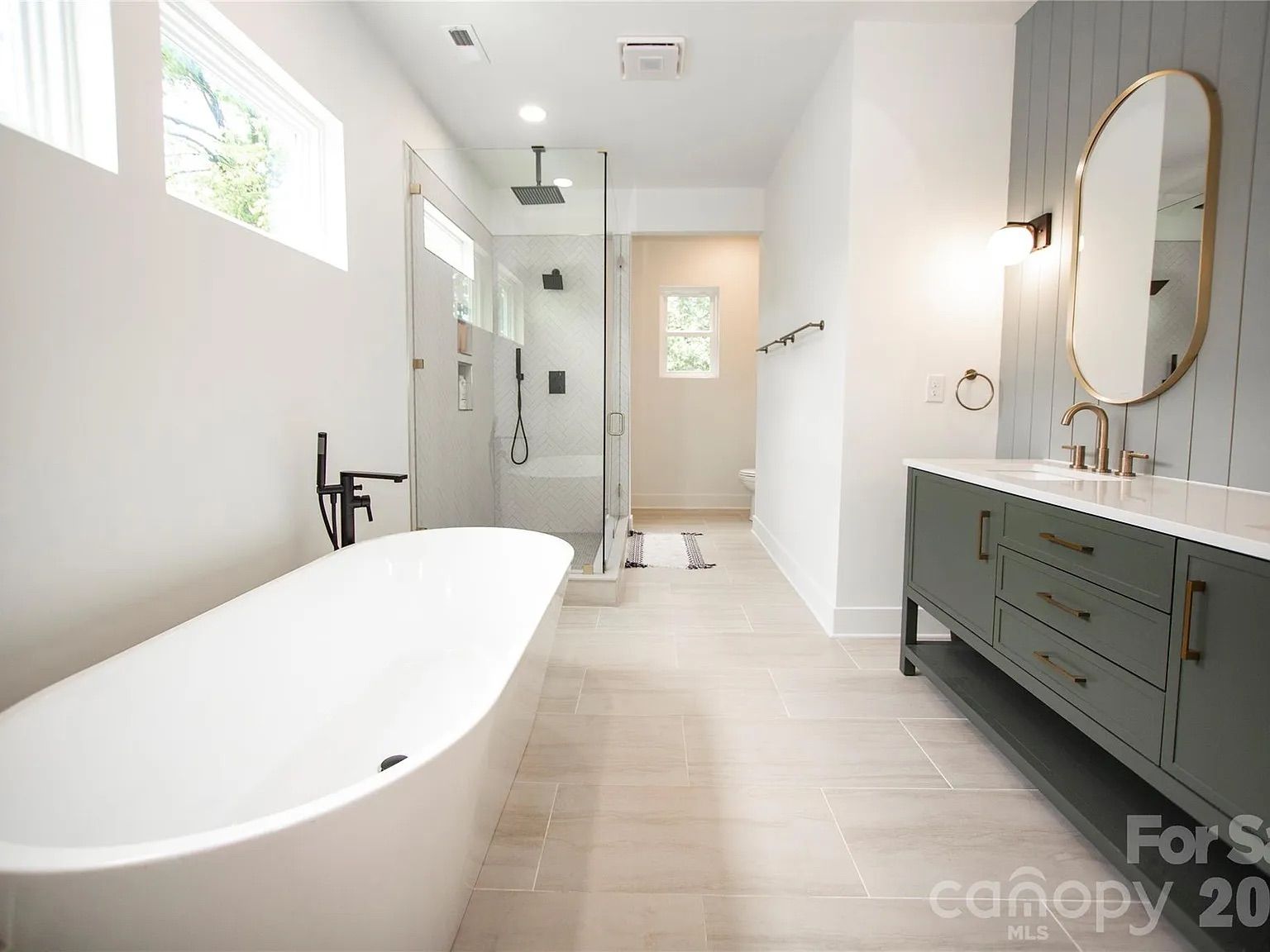 Bright modern bathroom with a white freestanding tub left, glass shower in center, and a gray vanity with brass fixtures on the right; pale tile floor and large mirror.