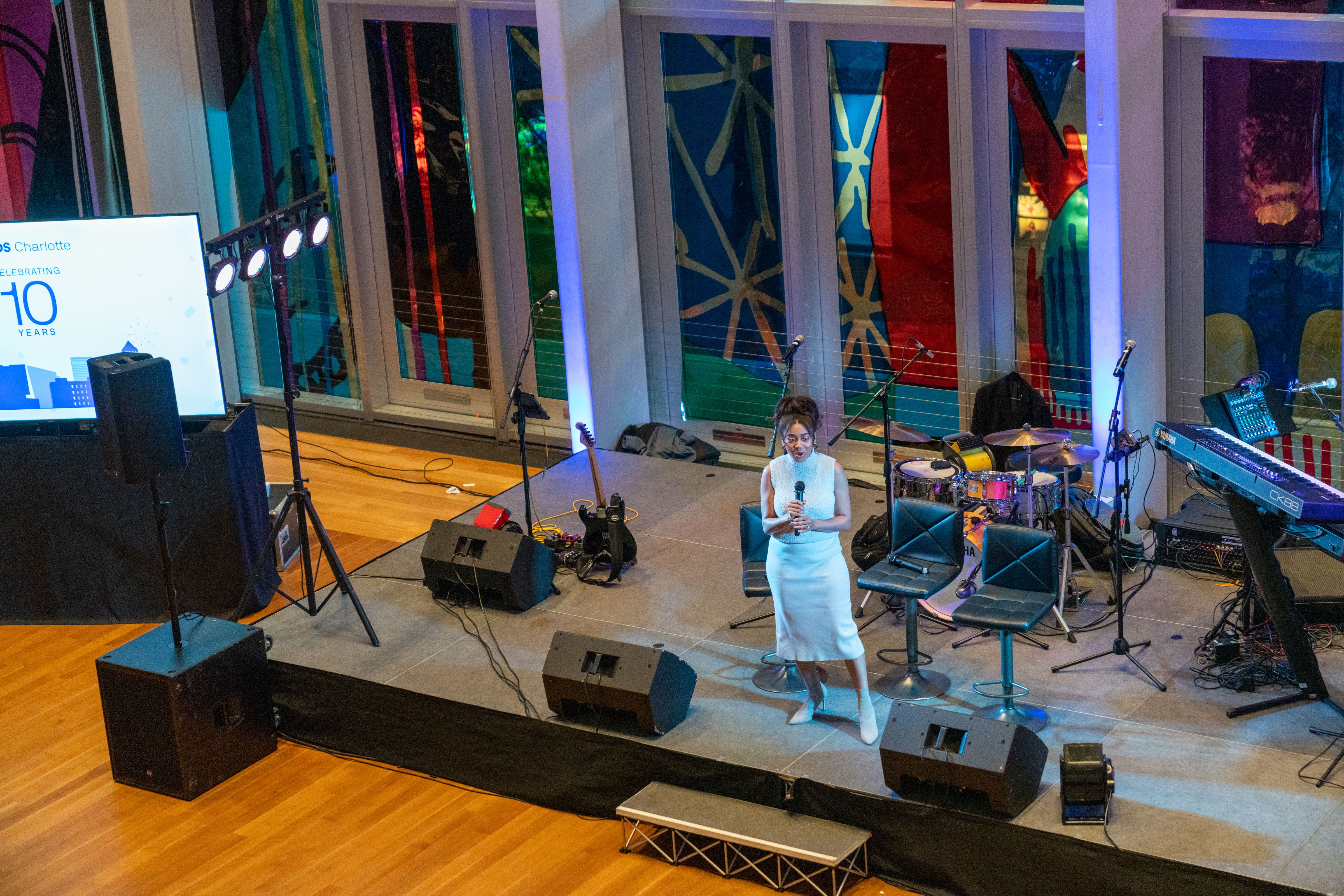 Woman in white dress holding microphone on stage with musical instruments and colorful abstract window panels in background during event.