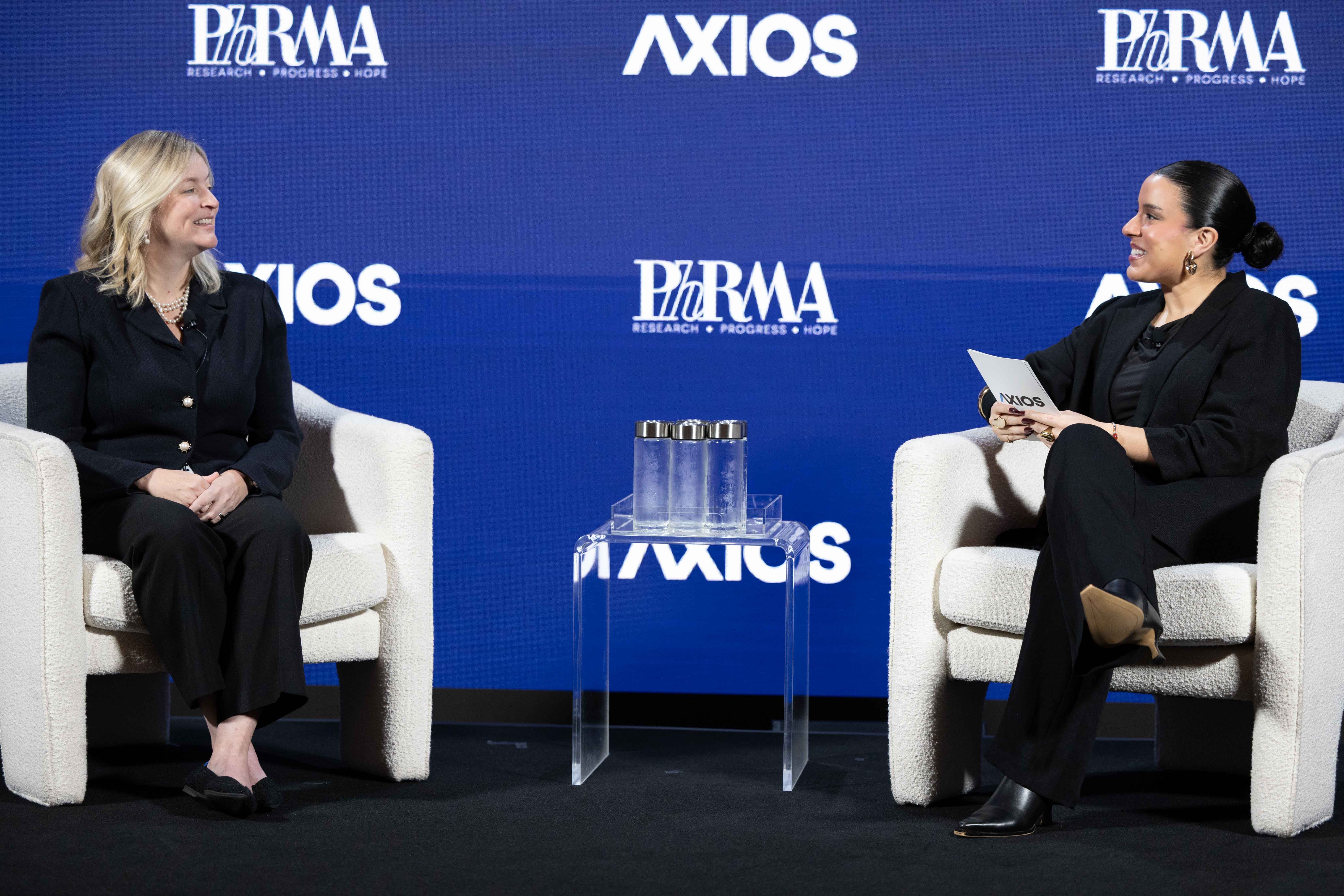 Two women sit on white armchairs on a blue backdrop with Axios logos. Left: blonde in a black suit with pearls; right: dark-haired woman in black, holding a card. A glass display sits between them.