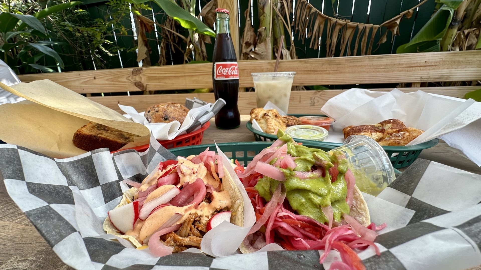 Two tacos with pickled onions, radishes and sauces in a basket with checkered paper on a wooden table outdoors, accompanied by a Coca-Cola bottle, iced drink and baskets of baked goods.