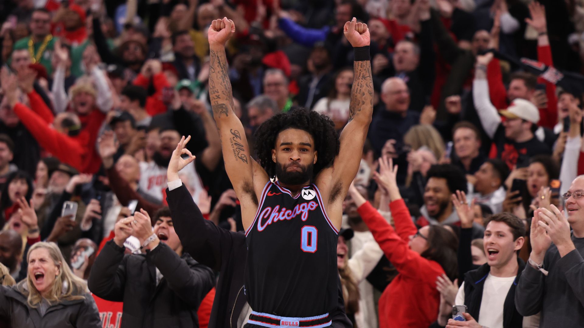 Chicago Bulls basketball player wearing black jersey with number 0 raises arms in celebration amid excited crowd cheering and clapping in a sports arena.