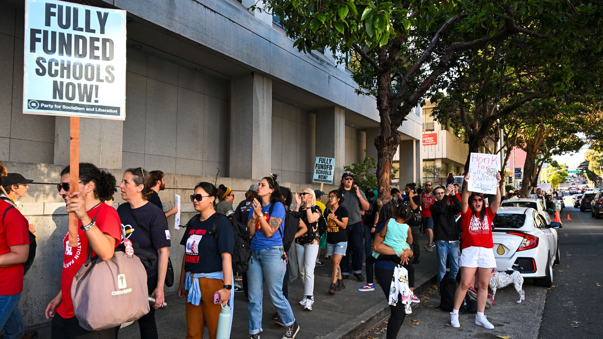 Protesters standing on a city sidewalk next to a building with trees, holding signs reading "FULLY FUNDED SCHOOLS NOW!" and "Honk for Fully Staffed Schools" in daytime.