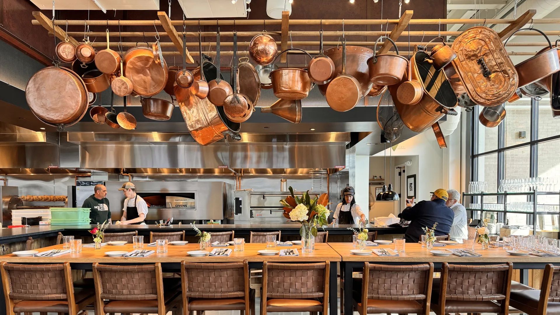 Modern restaurant kitchen with copper pots hanging overhead, wooden chairs and tables set with plates and glasses, chefs working, and two people seated by large windows.