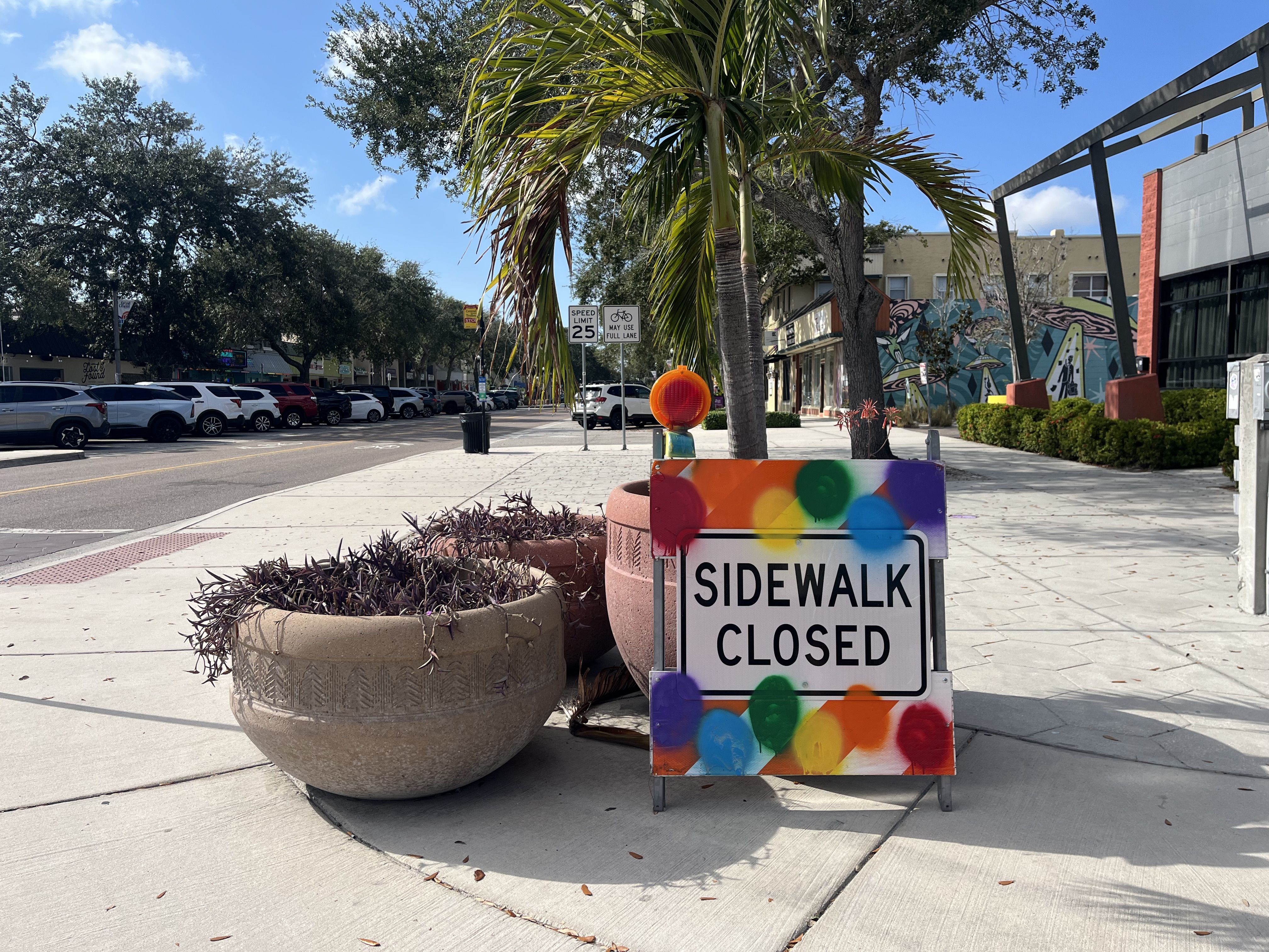 Rainbow-painted "SIDEWALK CLOSED" sign next to large planters and a palm tree on a sunny street with parked cars and buildings in the background under a blue sky.