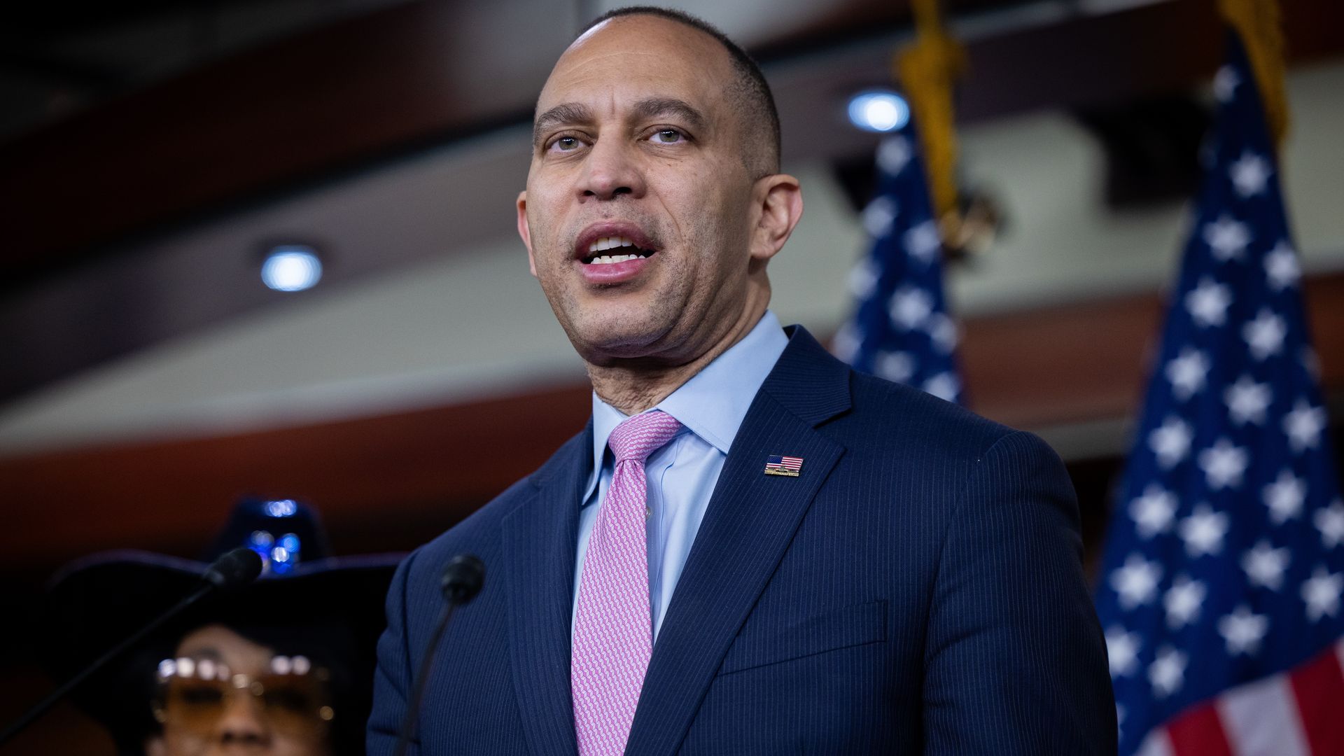 Man in a blue suit and pink tie speaking at a podium with two American flags in the background. A woman with glasses and a hat stands slightly blurred behind him.